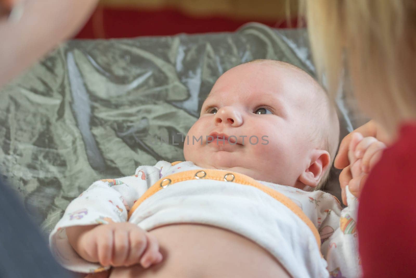 Top view of little baby lying on the blanket and looking at two women bending over him.