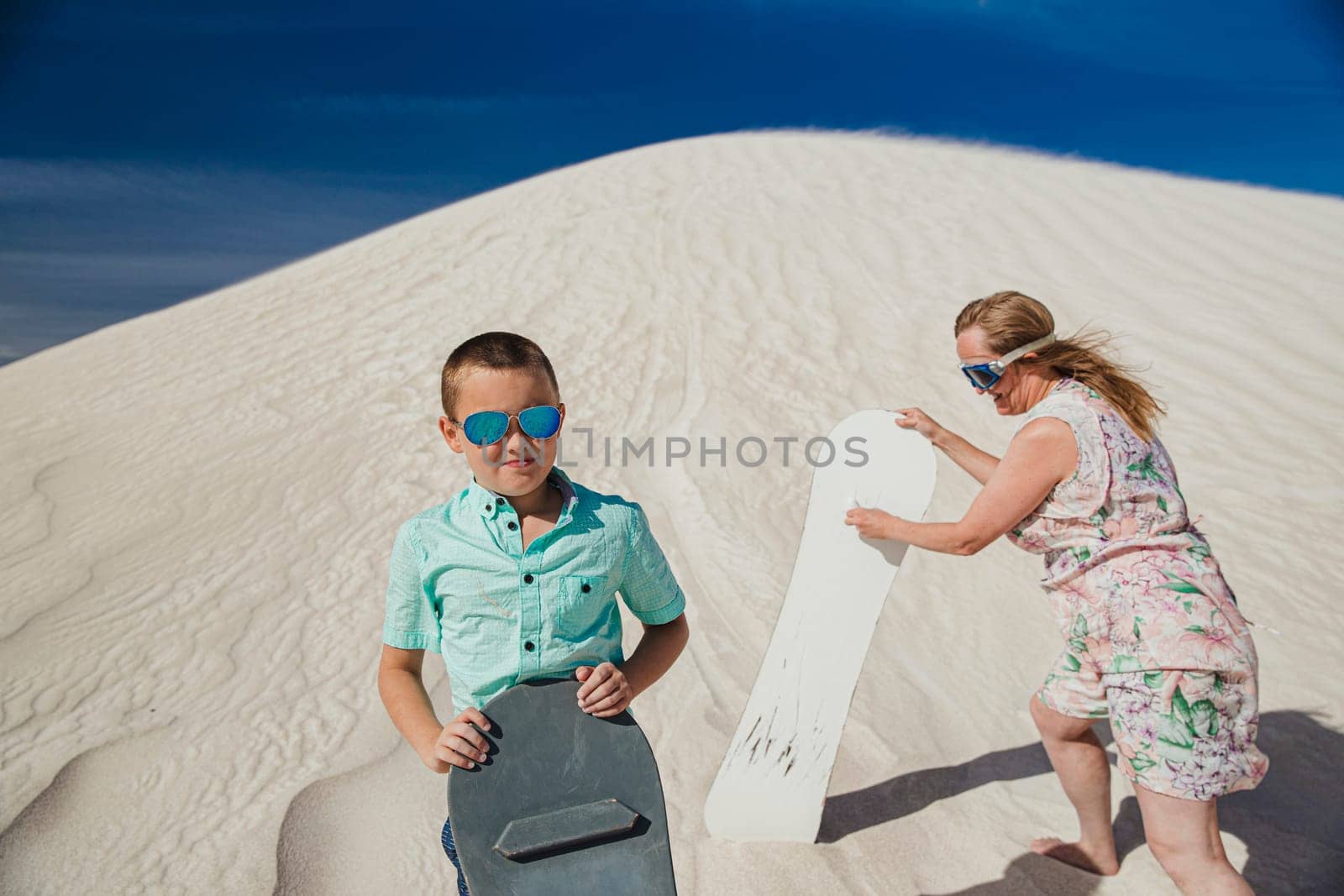 Portrait of a little boy stadning on a sand dune at Lancelin Sand Dunes. His mother is standing behind him, walking up the sand dune with a sand board.