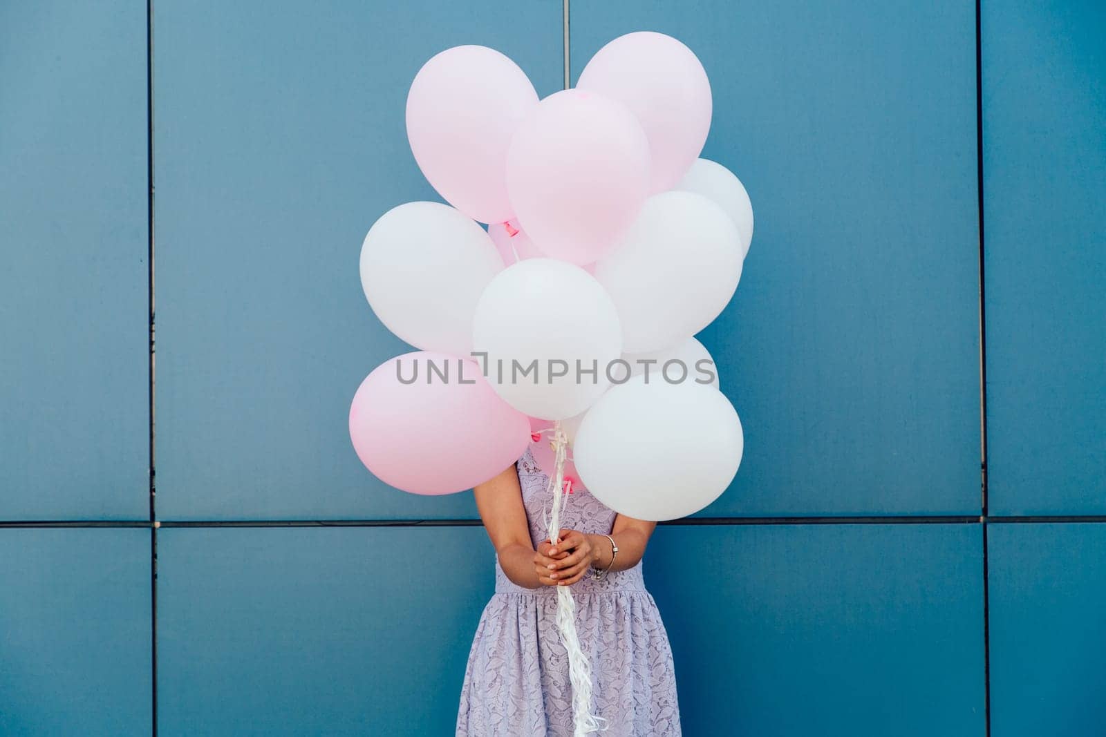 Young woman hiding her face with bunch of balloons, standing against blue wall. Dressed in tender dress. Outdoors.