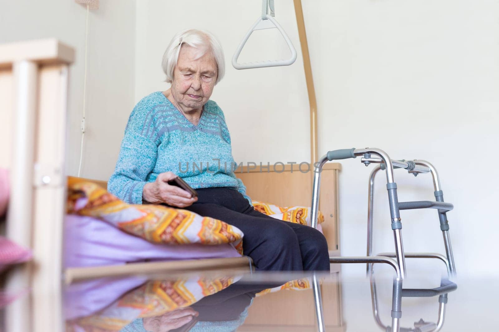 Elderly 96 years old woman reading phone message while sitting on medical bed supporting her by holder. Senior woman using wireless internet connection on smart phone.