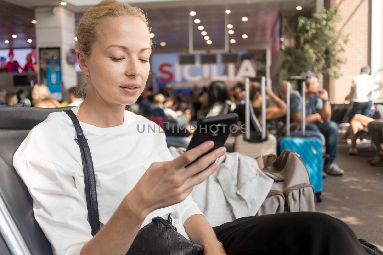 Casual blond young woman reading on her mobile phone while waiting to board a plane at the departure gates at the airport terminal. Travel concept.
