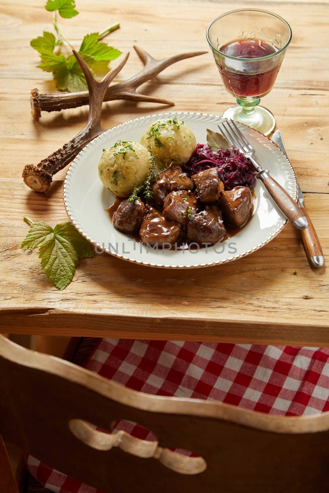 Plate of rich spicy wild venison goulash served with red cabbage and dumplings garnished with herbs on a rustic wooden table with red wine and deer antlers viewed over a chair