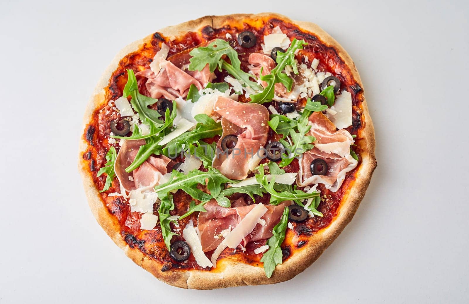 Tasty gourmet Italian pizza with prosciutto, black olives, parmesan cheese and fresh rocket viewed uncut from overhead on white