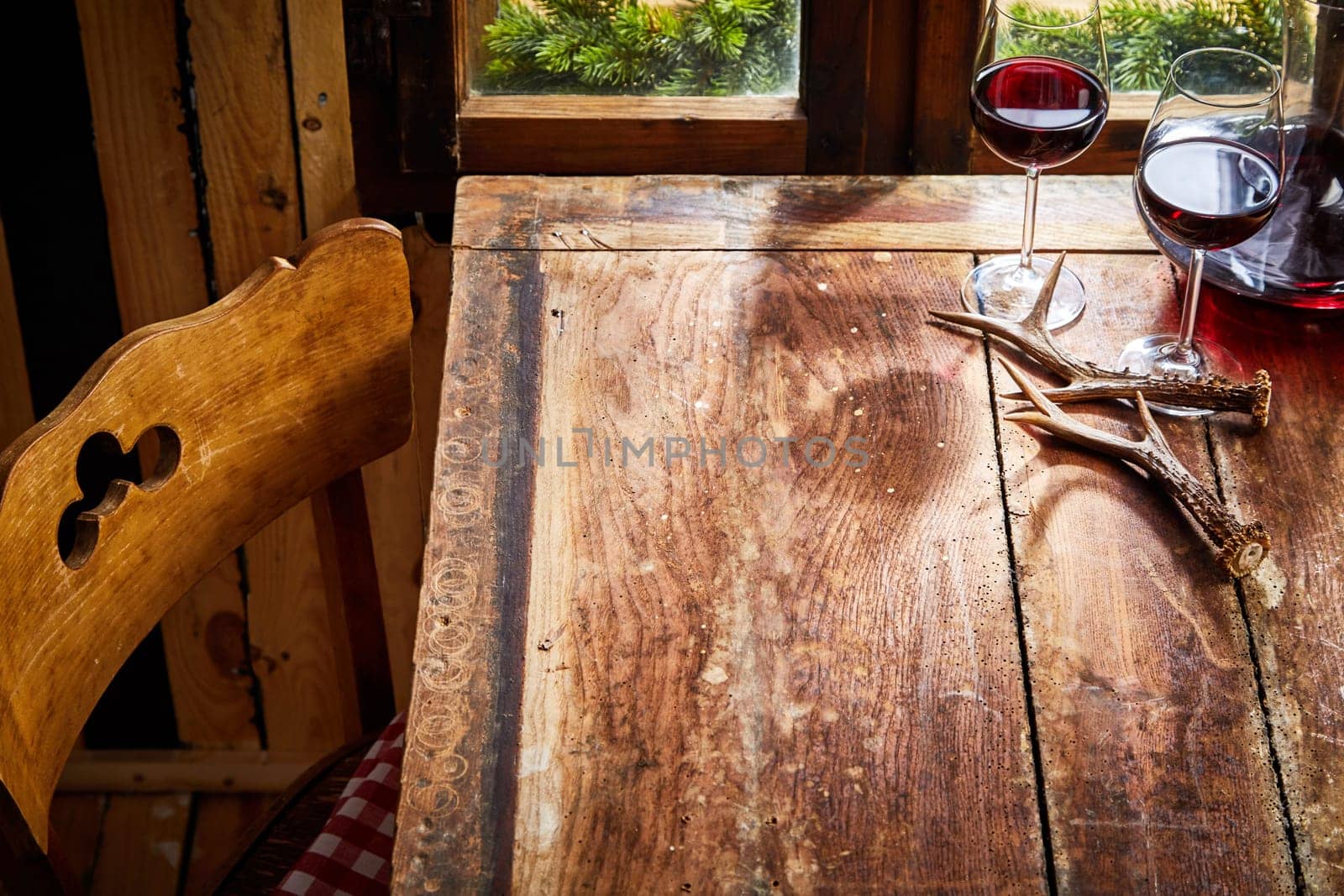 Old bare rustic wooden chair and table with deer antlers and two glasses of red wine in front of the window for food or product placement