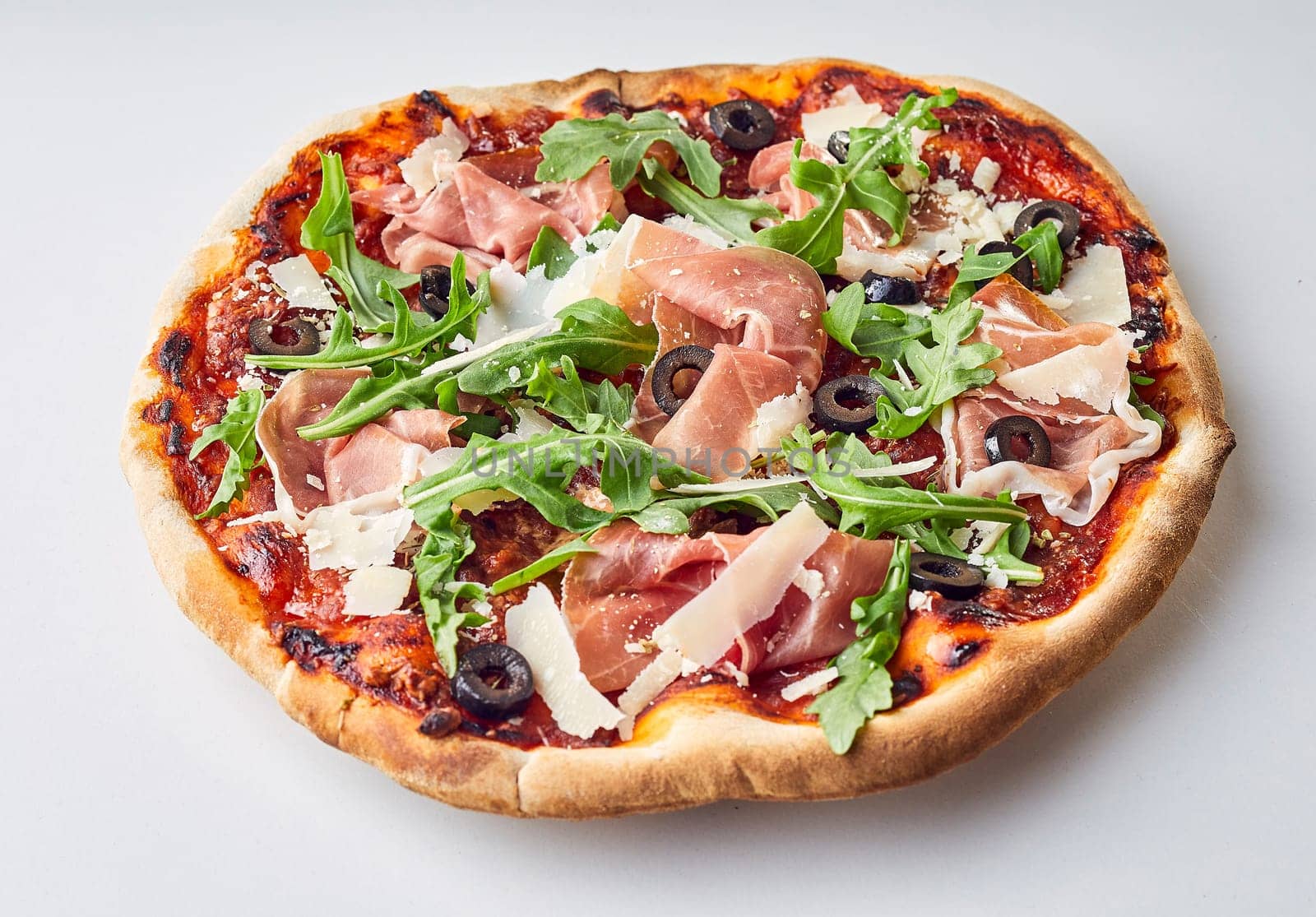 Traditional flame grilled crispy Italian pizza on a thin base topped with prosciutto, black olives, parmesan cheese and fresh rocket over a white background