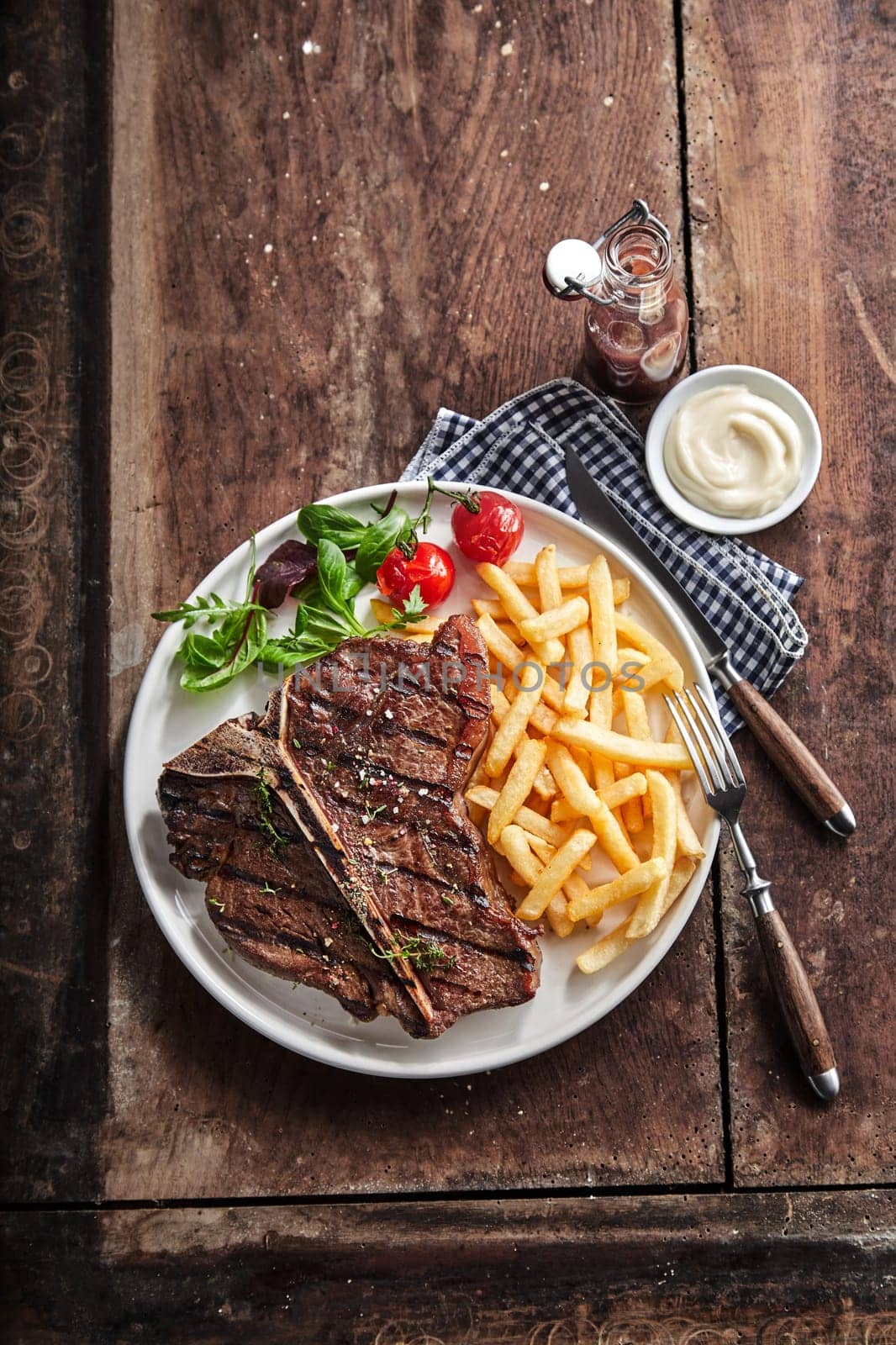 Grilled lean T-bone steak with golden potato chips and salad served with a dip or dressing on a rustic wooden table in a top down view with copy space