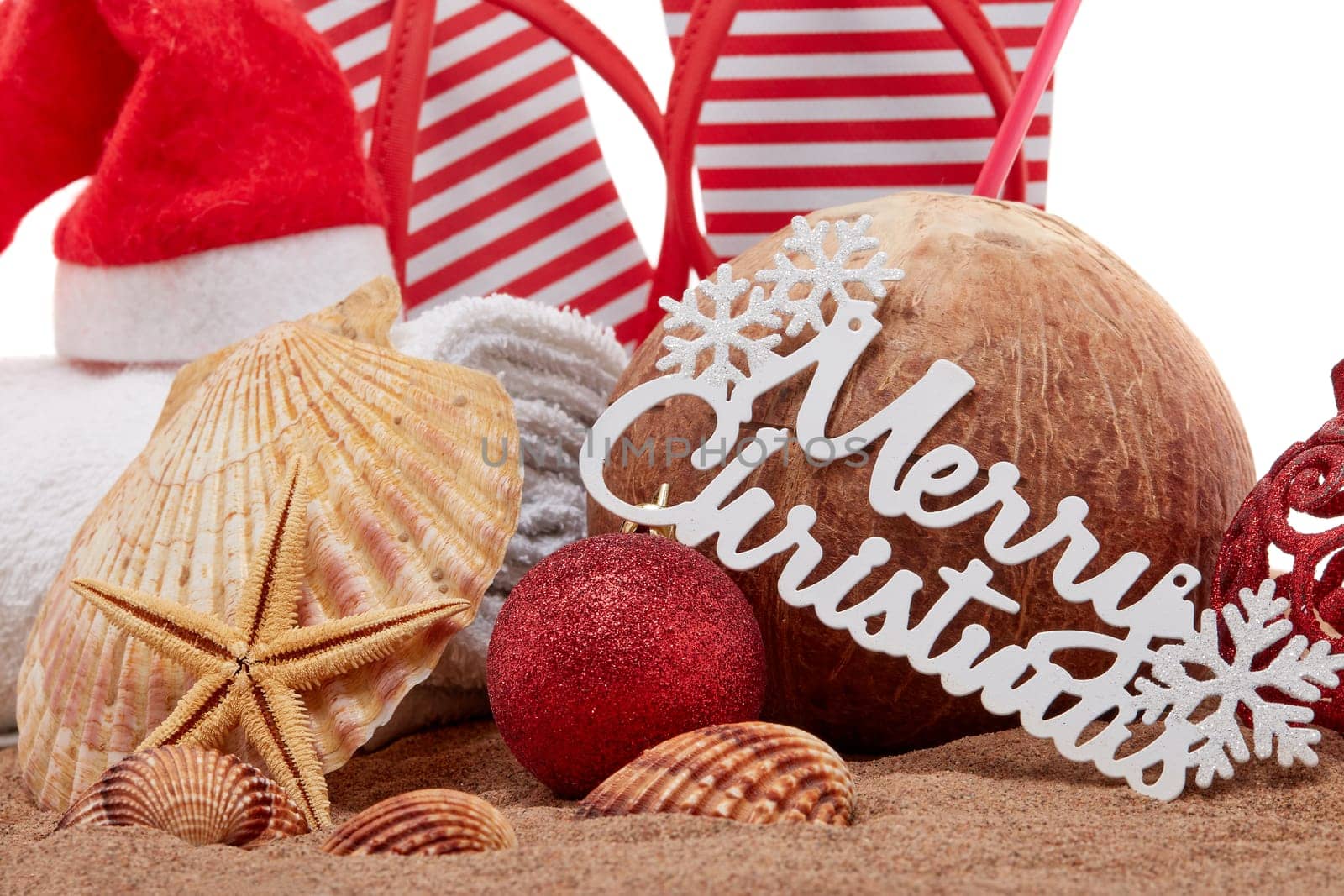 Still life with beach accessories (flip flops, towel, seashells, sand, coconut) and christmas ornaments on white background. Christmas. Winter vacation in warm countries, beach holiday