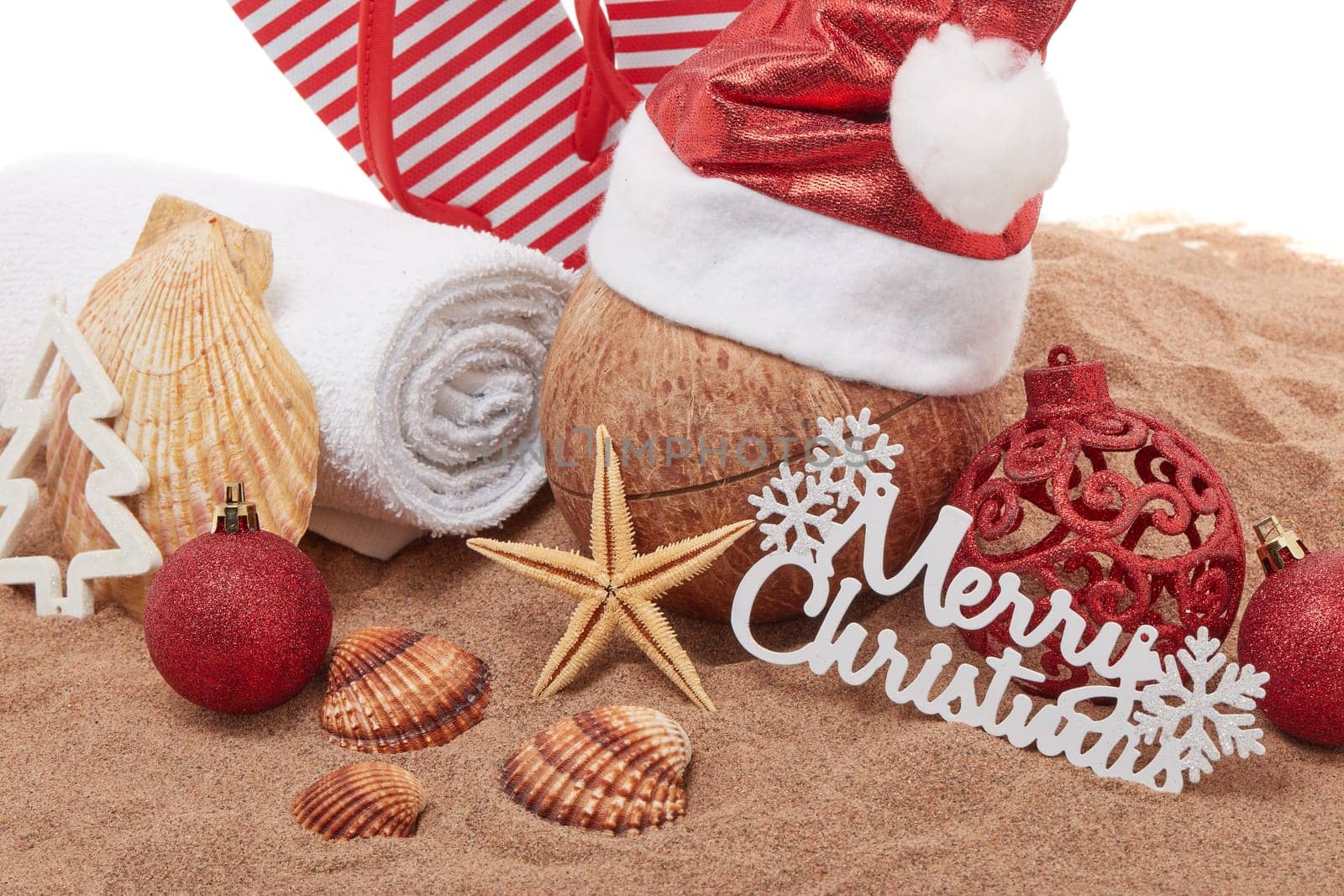 Still life with beach accessories (flip flops, towel, seashells, sand, coconut) and christmas ornaments on white background. Christmas. Winter vacation in warm countries, beach holiday