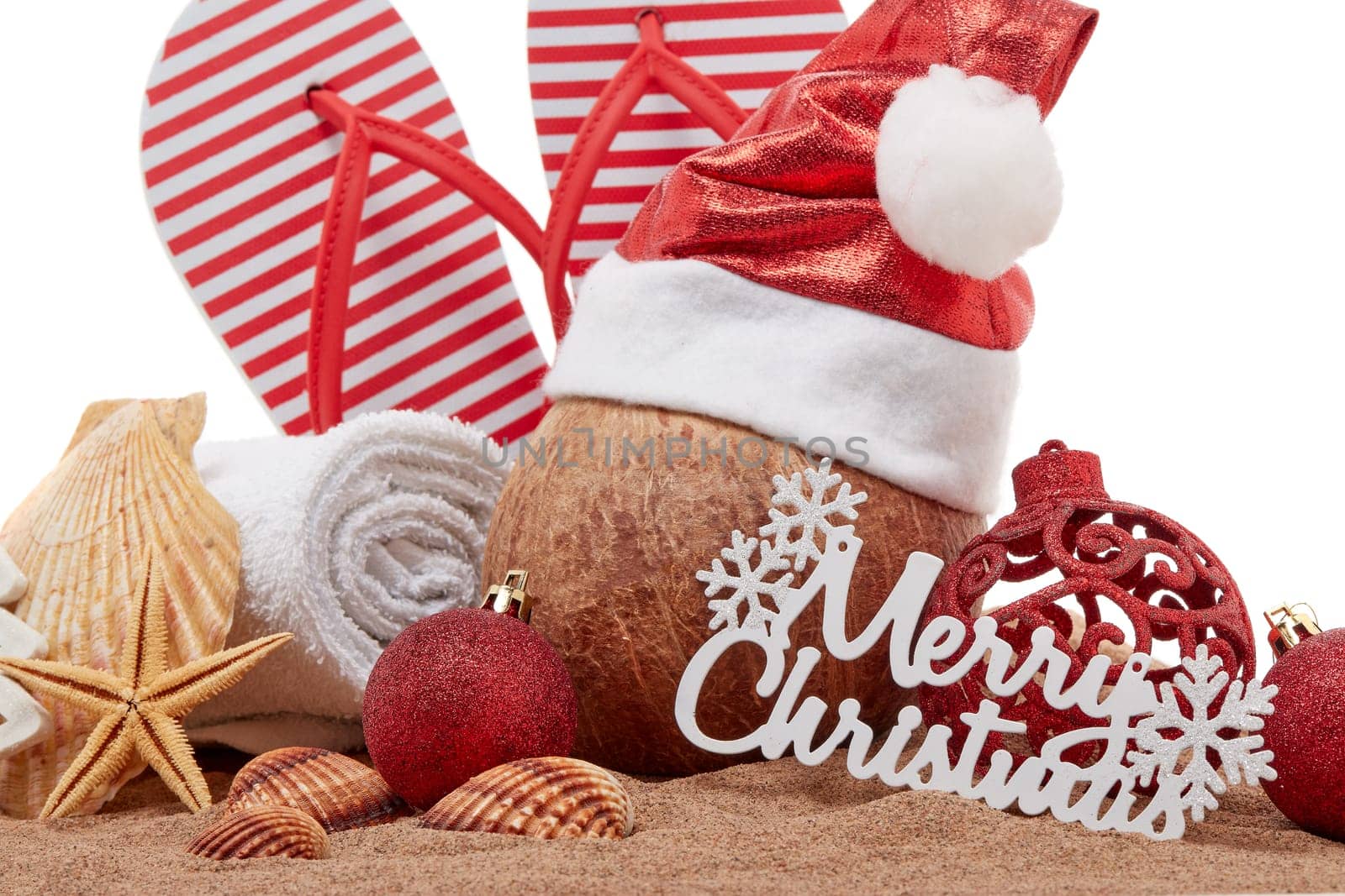 Still life with beach accessories (flip flops, towel, seashells, sand, coconut) and christmas ornaments on white background. Christmas. Winter vacation in warm countries, beach holiday