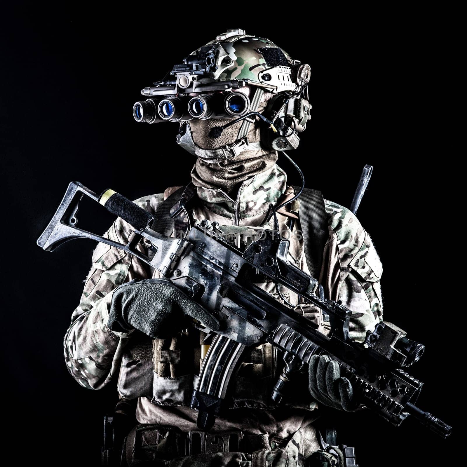 Marine rider in camouflage uniform and face mask, patrolling in darkness with quad-tube night vision goggles on battle helmet, holding modern assault rifle, low key studio portrait on black background
