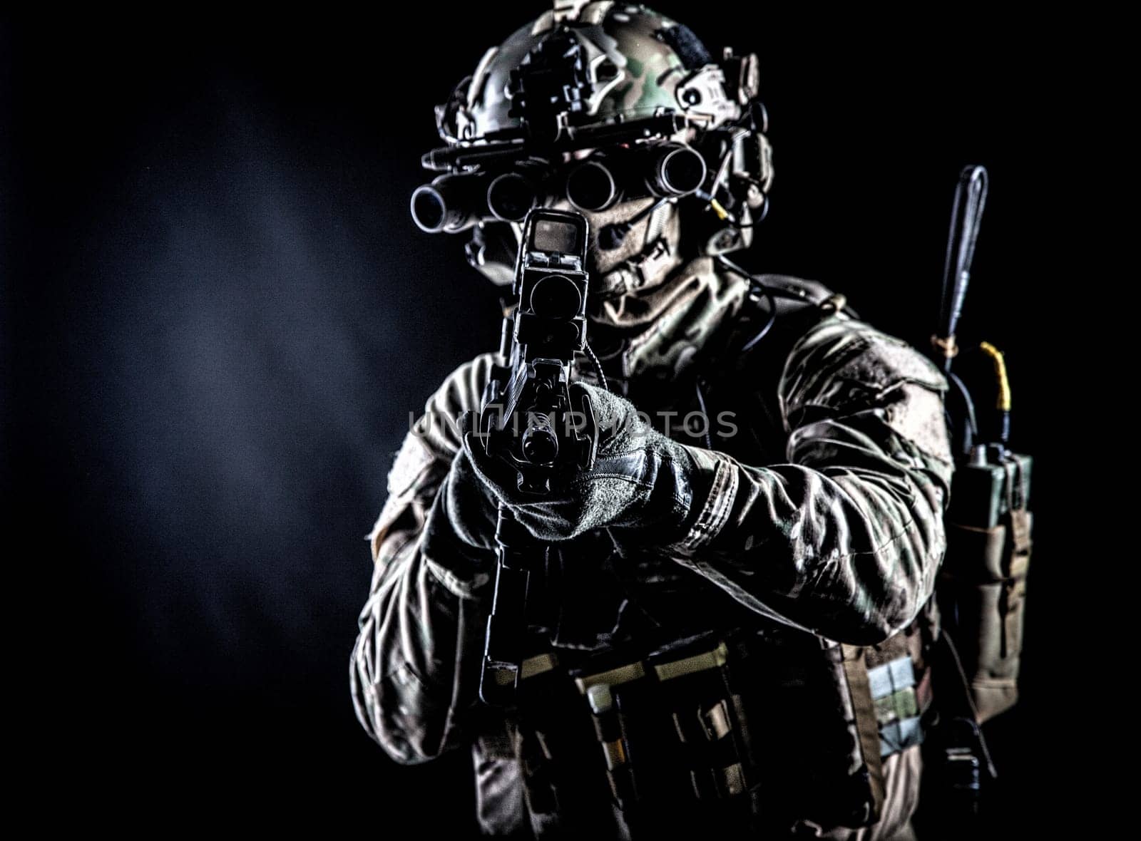 Soldier of special forces in camo combat uniform, load carrier, helmet, equipped night sight goggles, tactical radio headset, aiming assault rifle with collimator sight in camera, low key studio shoot