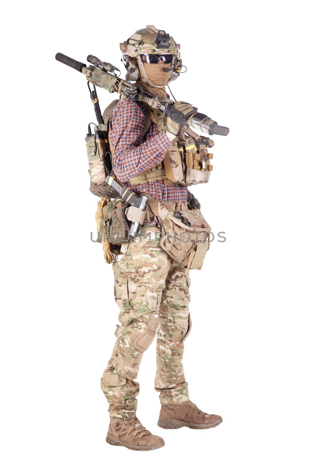Strikeball enthusiast in checkered shirt wearing military ammunition, face mask, helmet and radio headset, tactical glasses, camo pants, armed service rifle and handgun studio shoot isolated on white