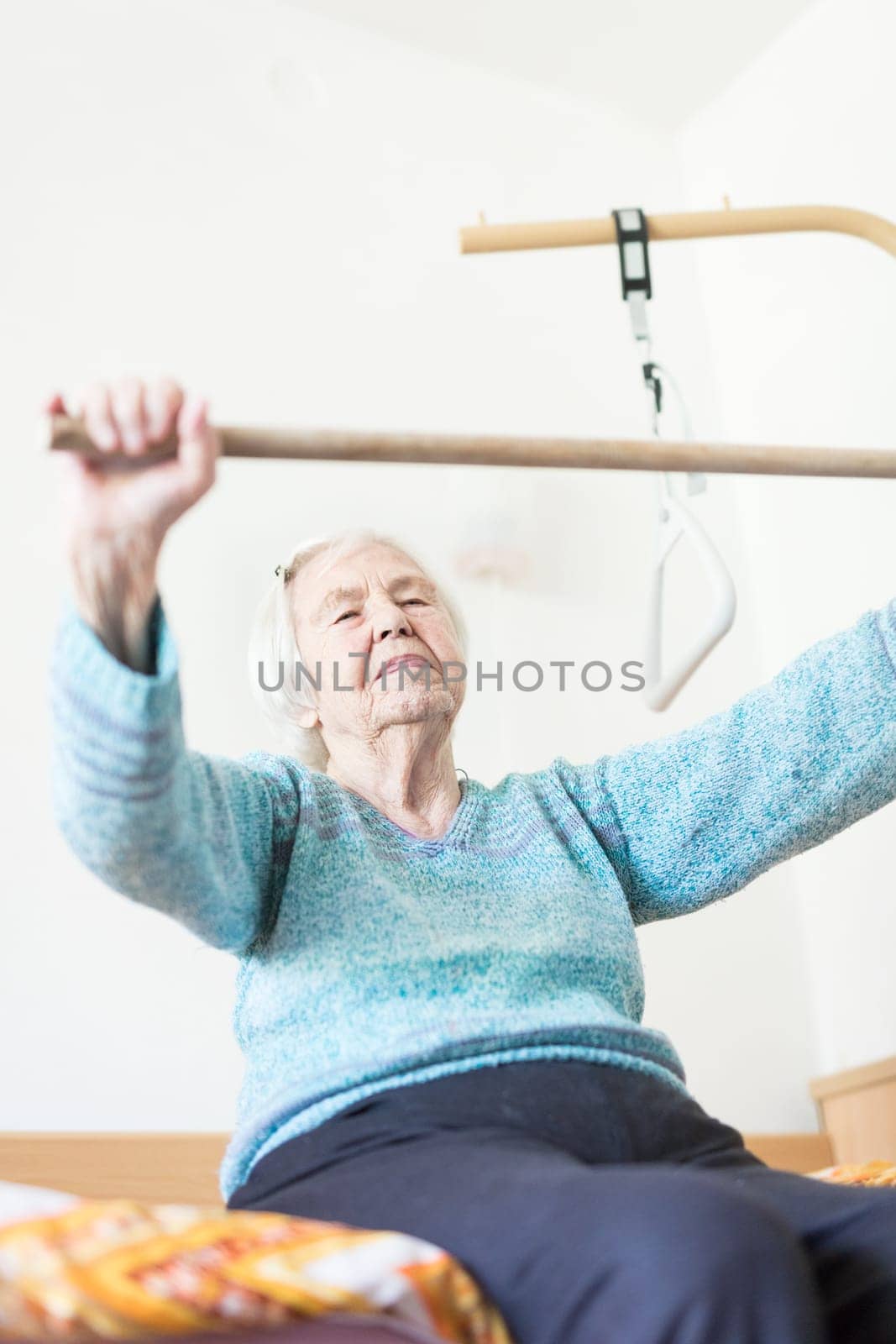 Elderly 96 years old woman exercising with a stick sitting on her bad. Geriatric health care home assisted support for older people concept. Care for the elderly.