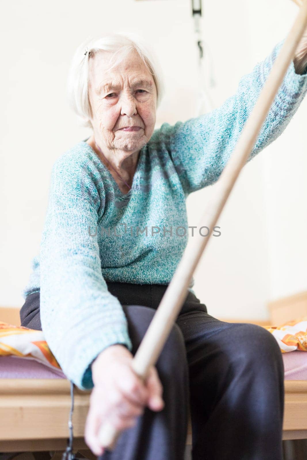 Elderly 96 years old woman exercising with a stick sitting on her bad. Geriatric health care home assisted support for older people concept. Care for the elderly.