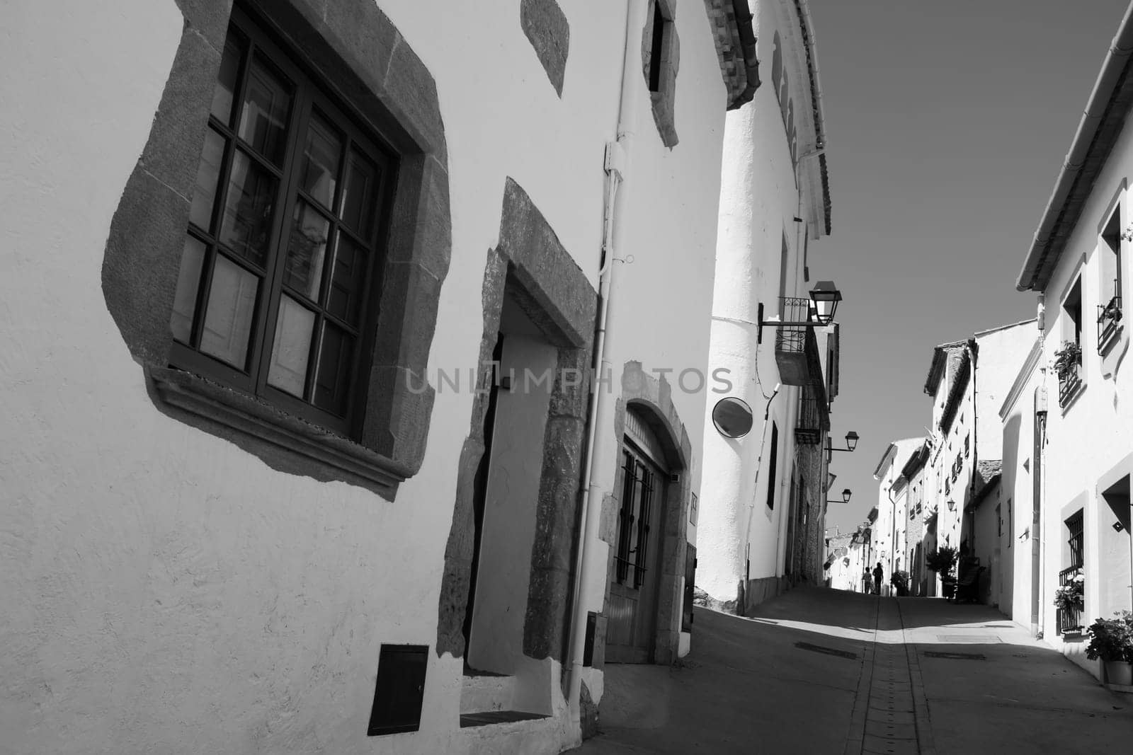 Low angle view of white houses along a narrow street in Begur. Two children walk away in the distance. Black and white wide-angle photo.