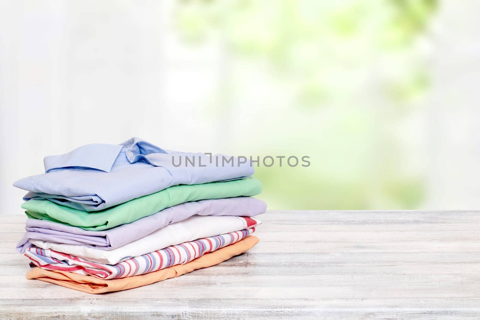 Stack colorful clothes. Pile of folded cotton shirts on a bright table with space for your display product montage against abstract blurred natural light green background. Summer fashion.
