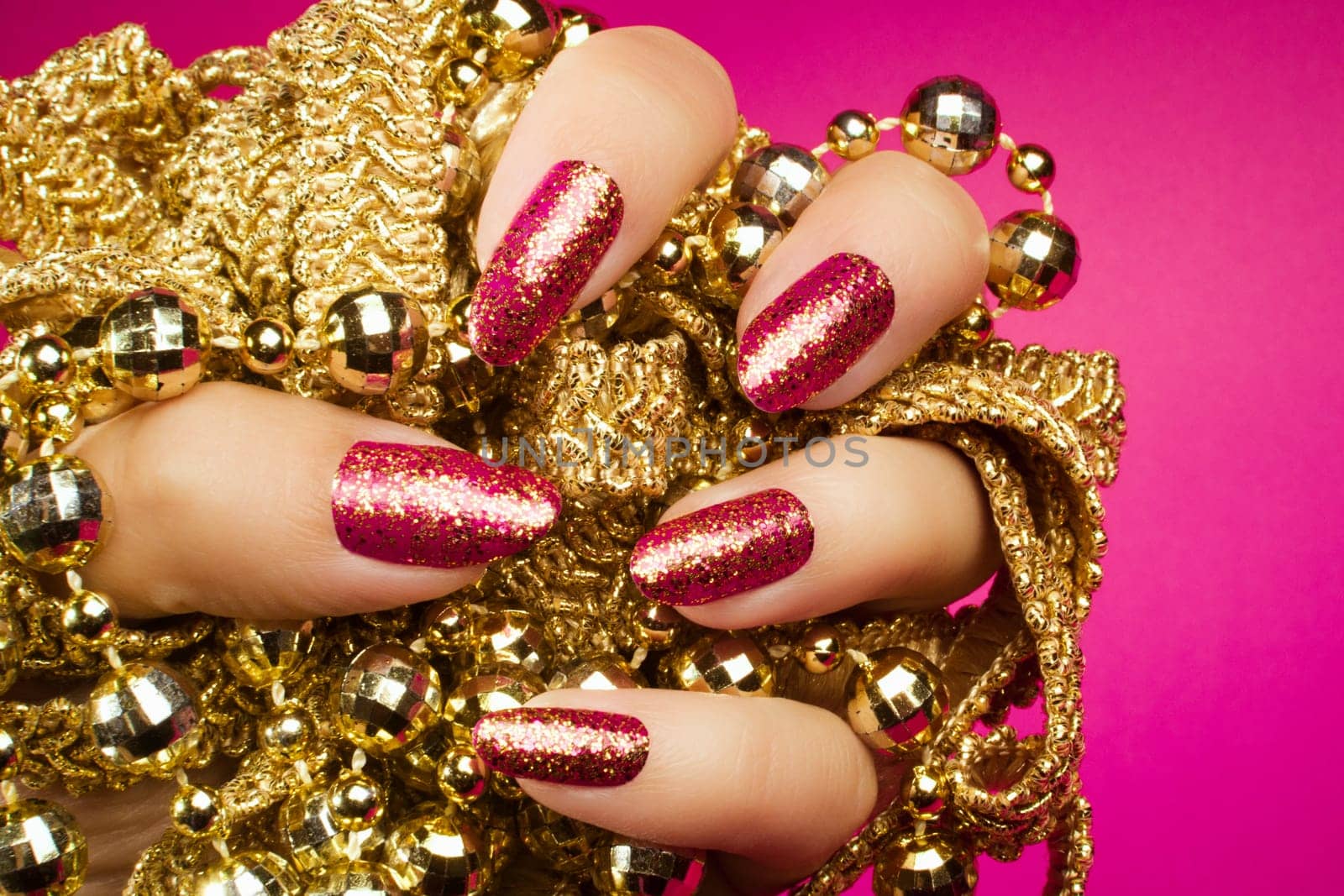 Female hand with gold glittered pink nails is holding gold pearl jewelry on pink background, nail care and manicure concept.