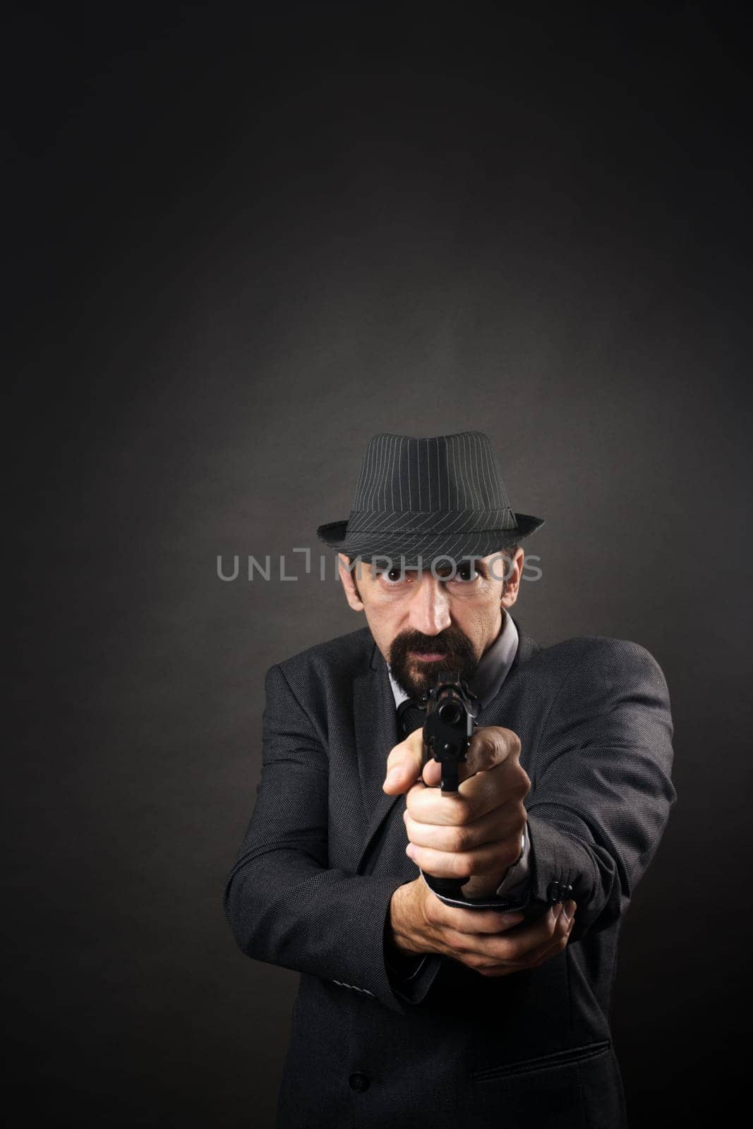 Old-fashioned gangster in suit and hat is aiming with pistol on dark background. Retro and vintage style.