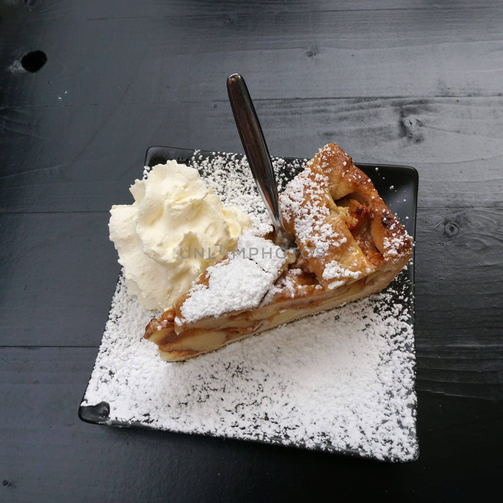 a piece of apple pie with cream and powdered sugar on a black, quatrefoil plate