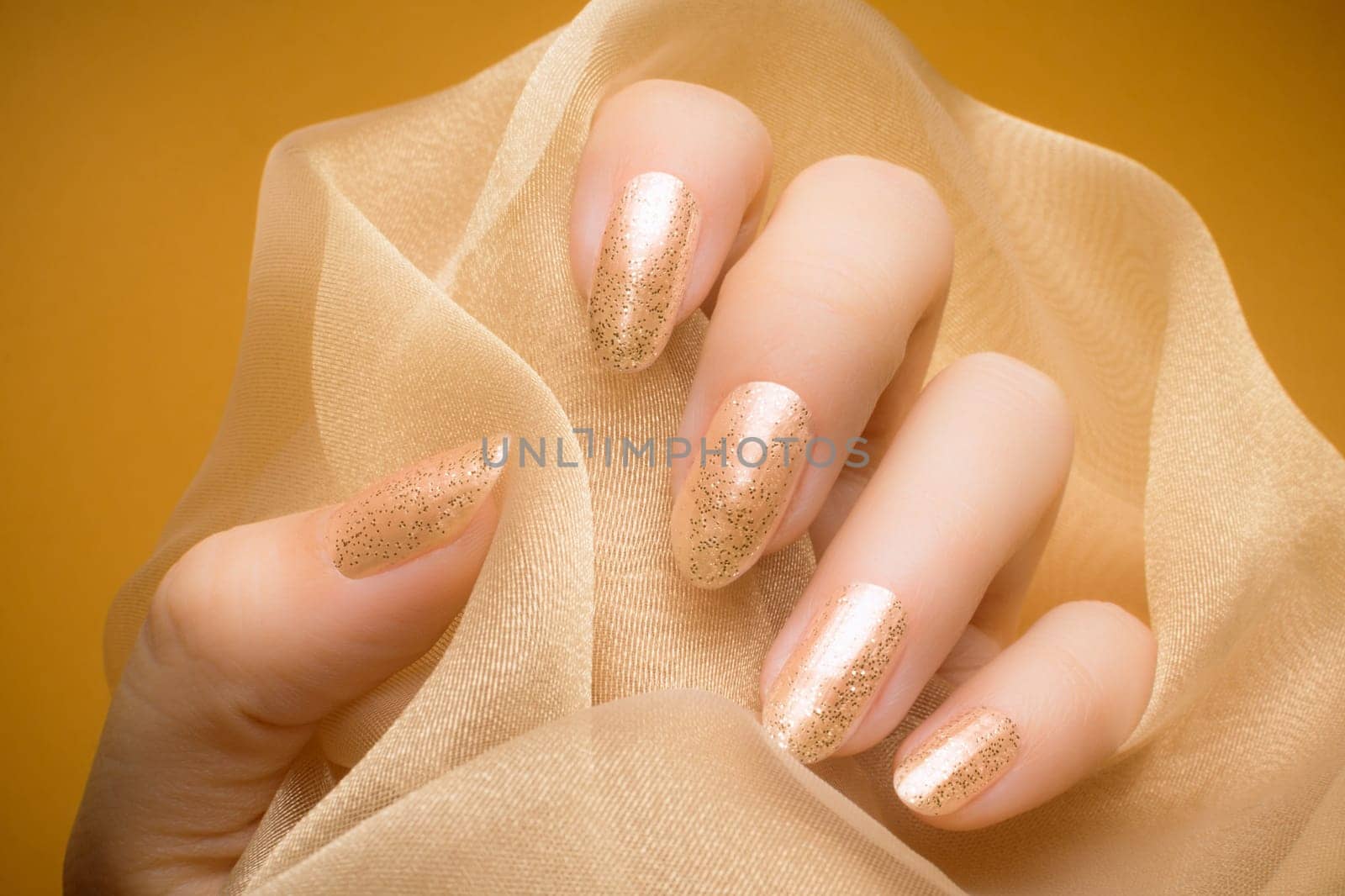 Female hand with glittered beige nails is holding beige textile on orange background.