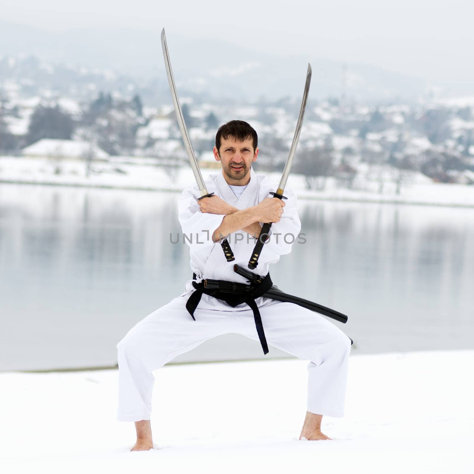 Man in white kimono and black belt is practicing martial arts with two samurai swords barefoot on snow at winter.