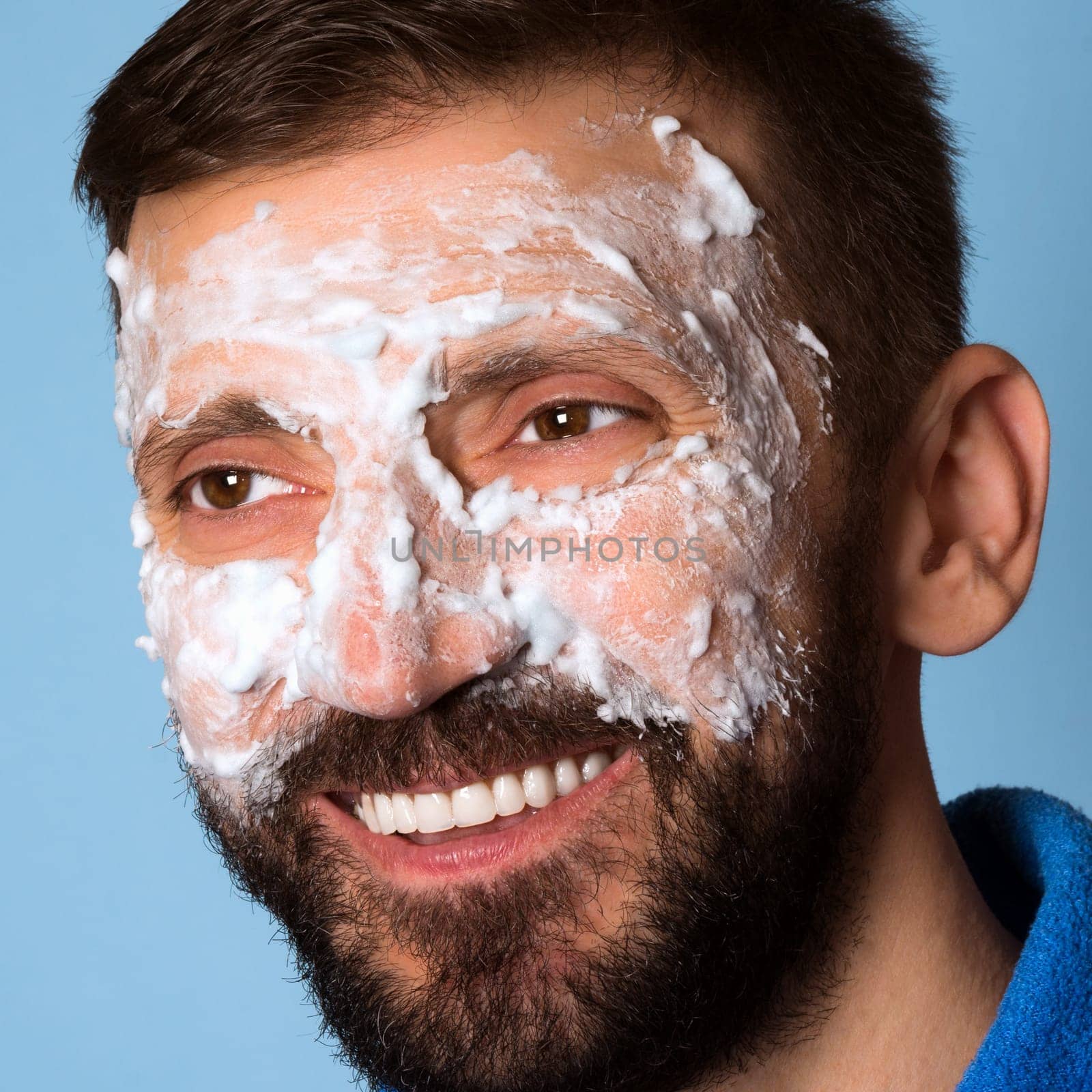 The smiling middle aged man is using foam for skincare on his face on blue background.