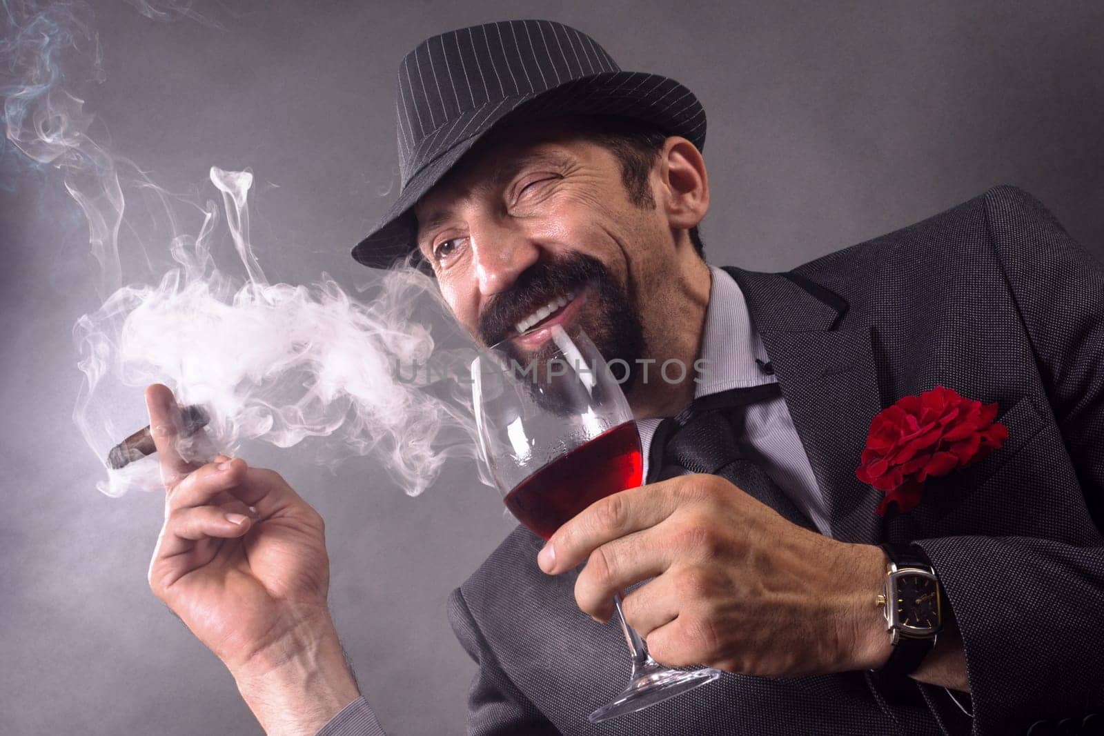 The smiling winking elegant gentleman in suit and hat is smoking cigar and drinking wine on gray background.
