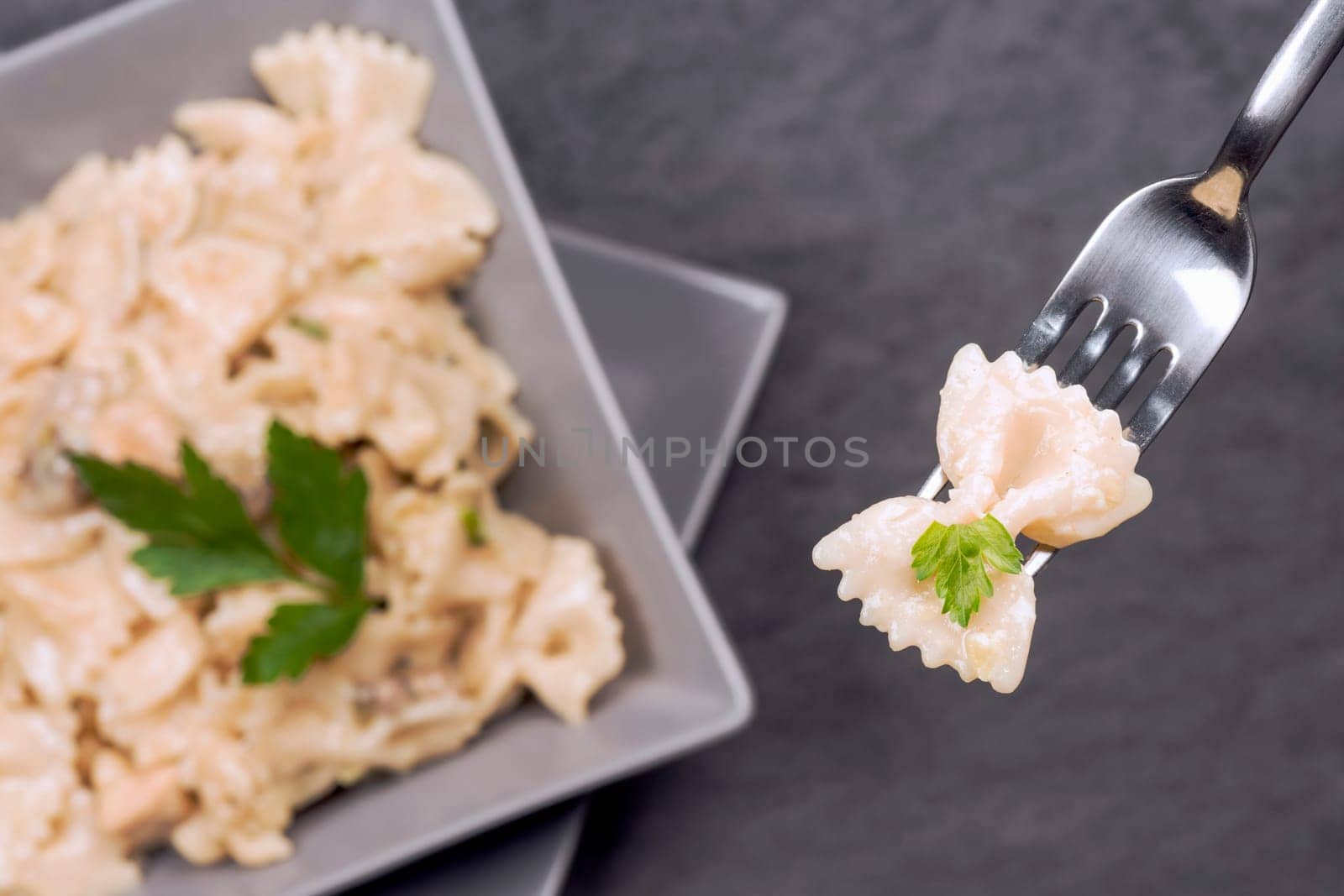 Pasta Italian food is on fork and in gray square plate on dark gray background.