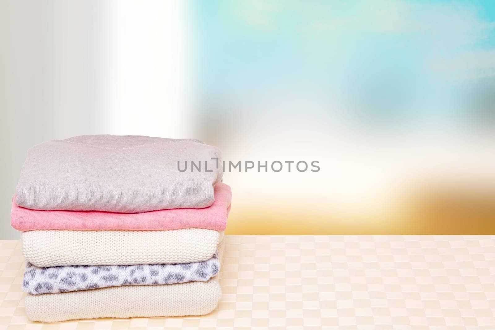 Stack of winter clothes. A pile of colorful cozy warm sweaters on a bright wooden table against abstract blurred natural background. Autumn and winter clothes. Space for display product montage.