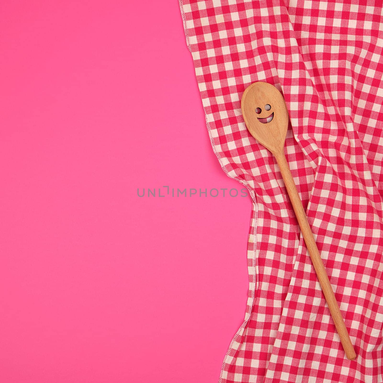 wooden spoon with a carved face on a red kitchen towel, pink background, copy space