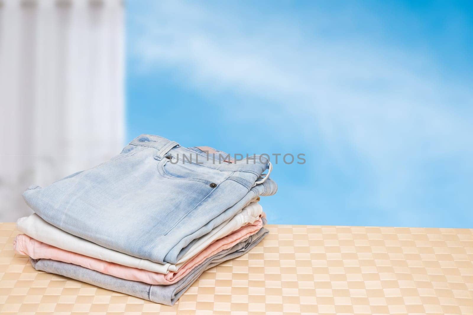 Stack of jeans. Closeup of a pile of colorful female denim pants on a bright table against abstract blurred cloudy sky. Space for your product display montage.