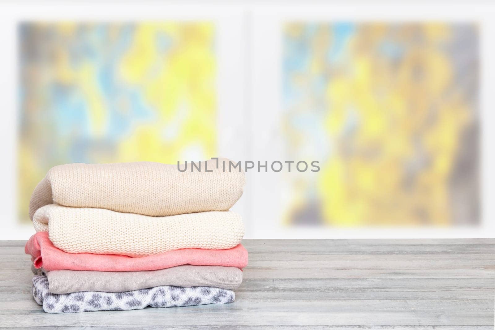 Stack of winter clothes. Pile of colorful knitted cozy warm sweaters or pullover on wooden table against blurred bright autumn background. Space for display product montage. Beautiful backdrop.