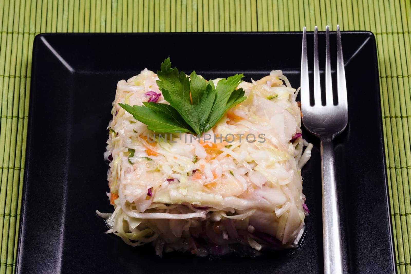 Mixed cabbage salad with fork is served on black square shape plate on green background.
