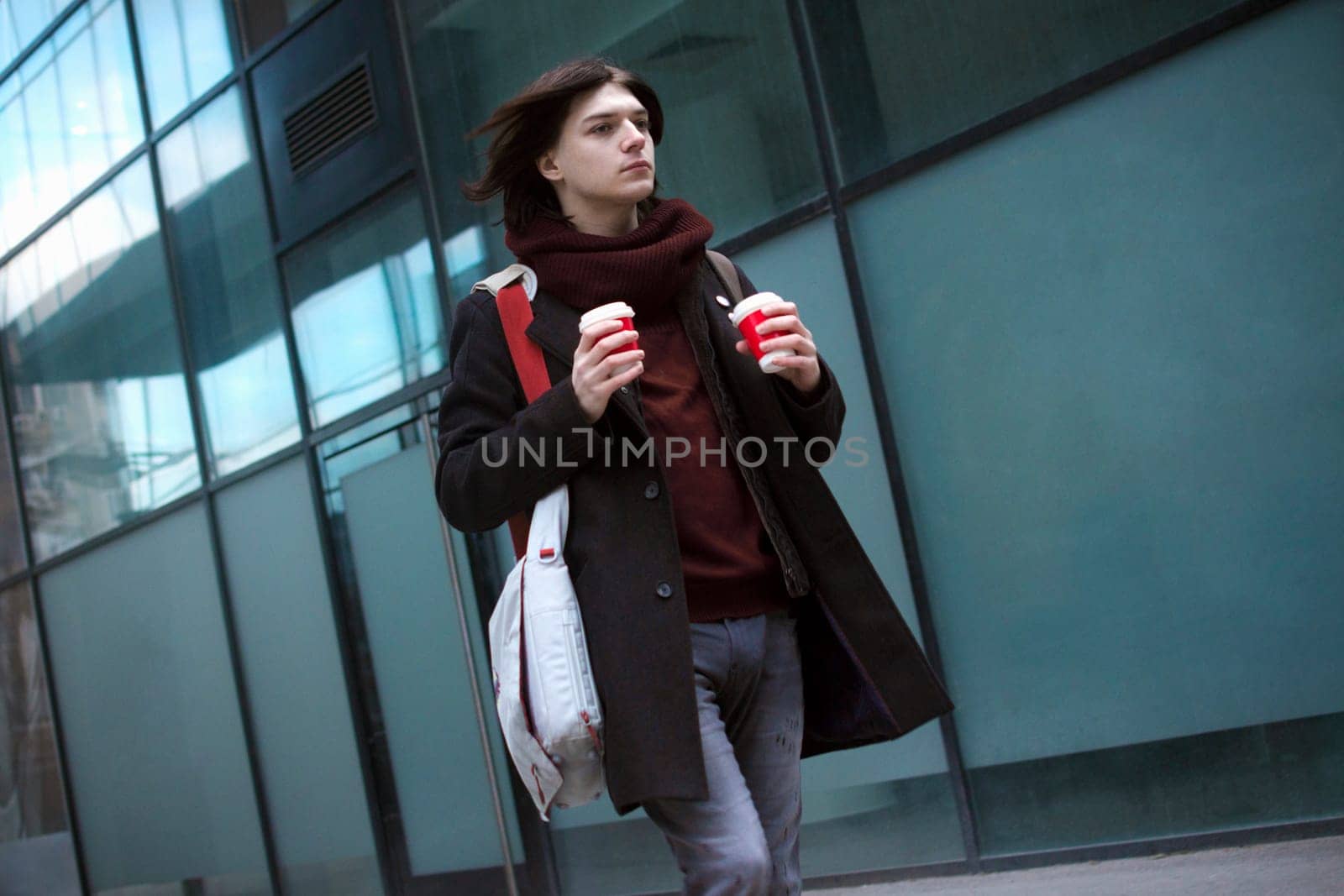 Trendy young guy with laptop bag and two coffees is walking or hurrying at the street. Work day morning.