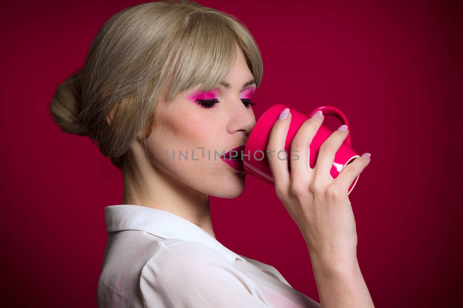 The fashionable trendy blond hair woman with pink make-up is drinking from red or pink mug on red background.