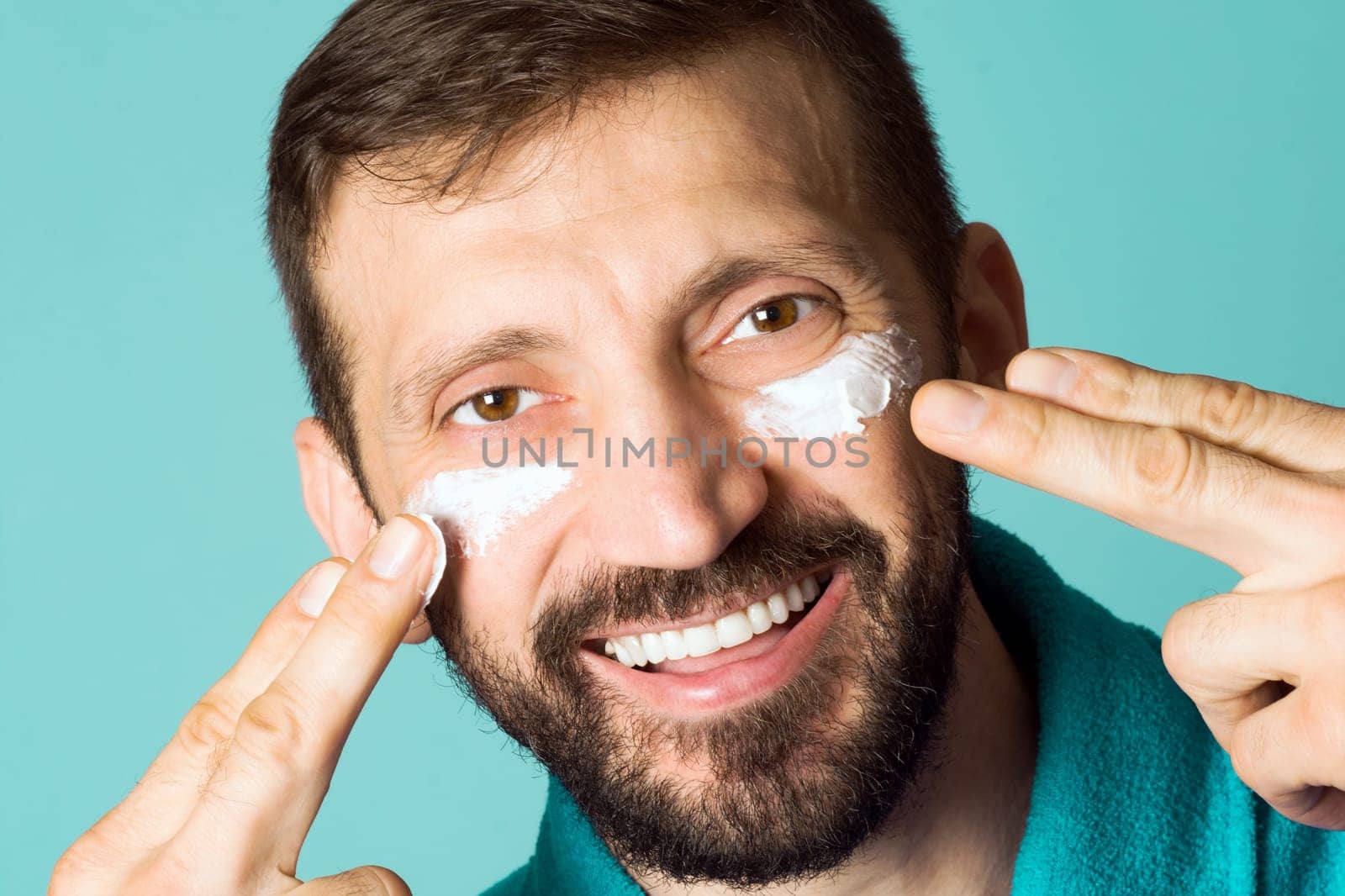 The smiling cheerful handsome man is applying face cream for men on his face on turquoise background.