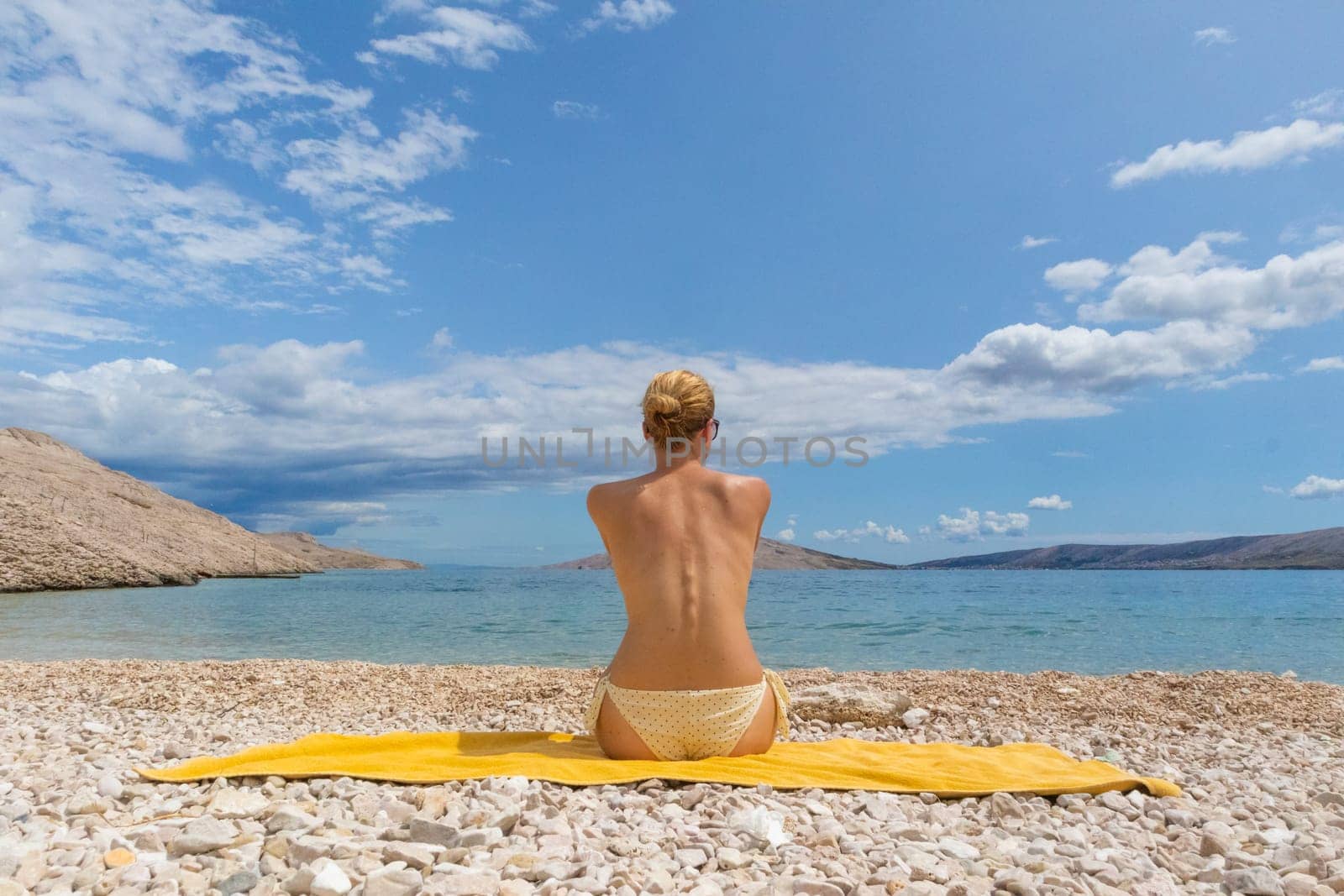 Rear view of relaxed sexy young caucasian woman sitting on yellow towel, sunbathing topless alone on romote pabble beach on Pag island, Croatia, Mediterranean. Enjoying pure nature.
