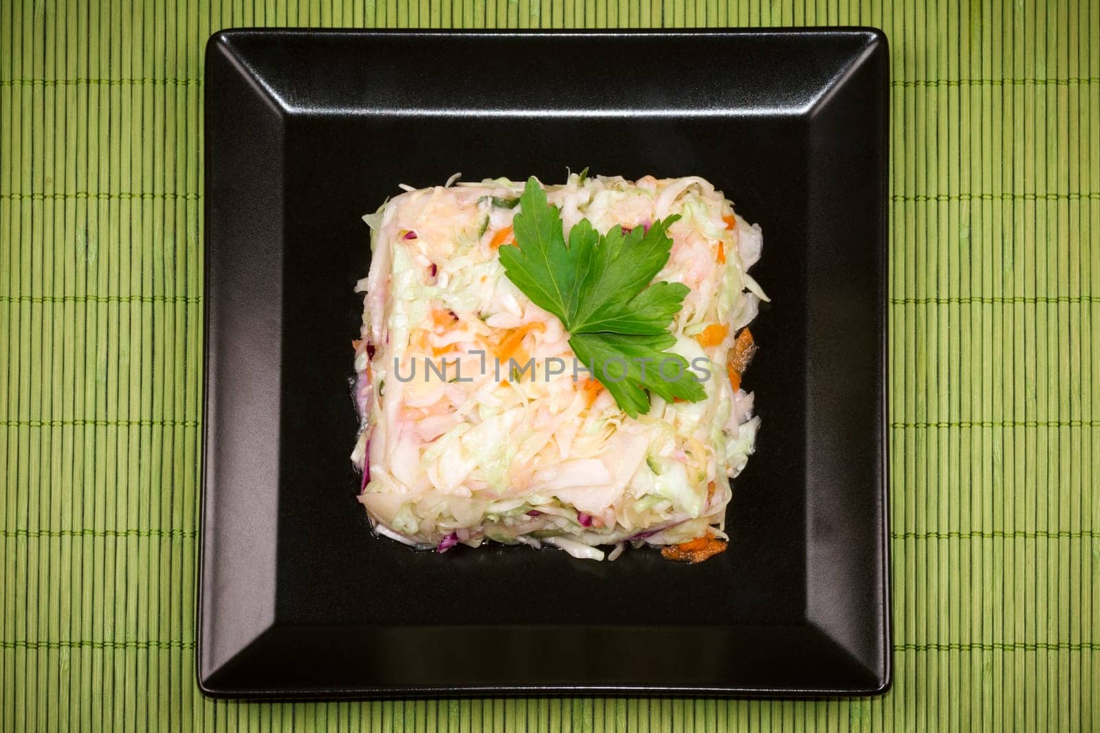 Mixed cabbage salad with parsley decoration is on black plate on green tablecloth.