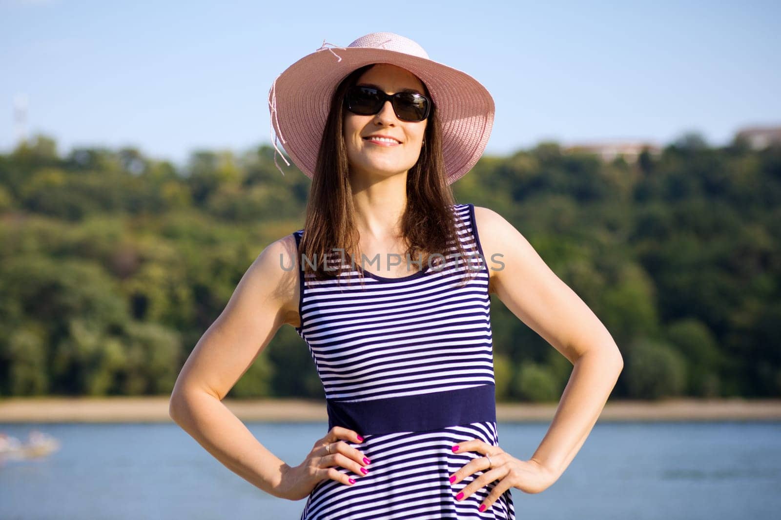 Attractive woman in hat, sunglasses and dress is enjoying in summer at the riverside.