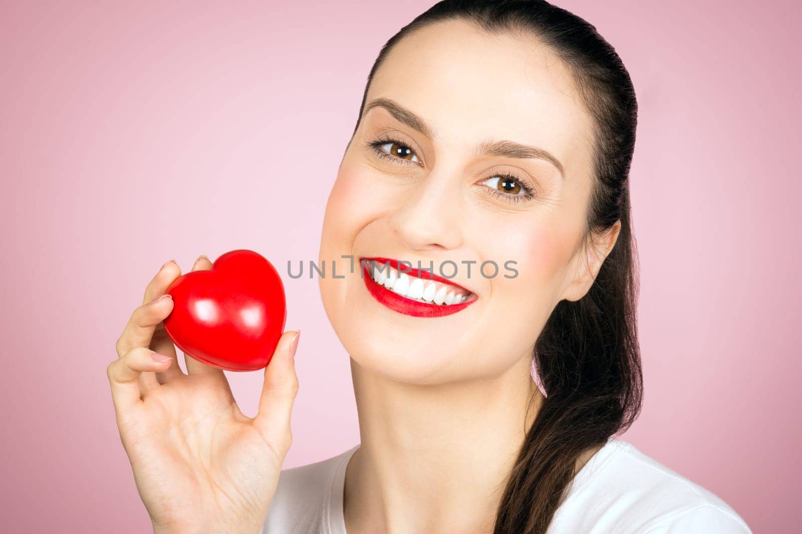 Beautiful smiling woman is holding a little red heart in hand on pink background, love concept.