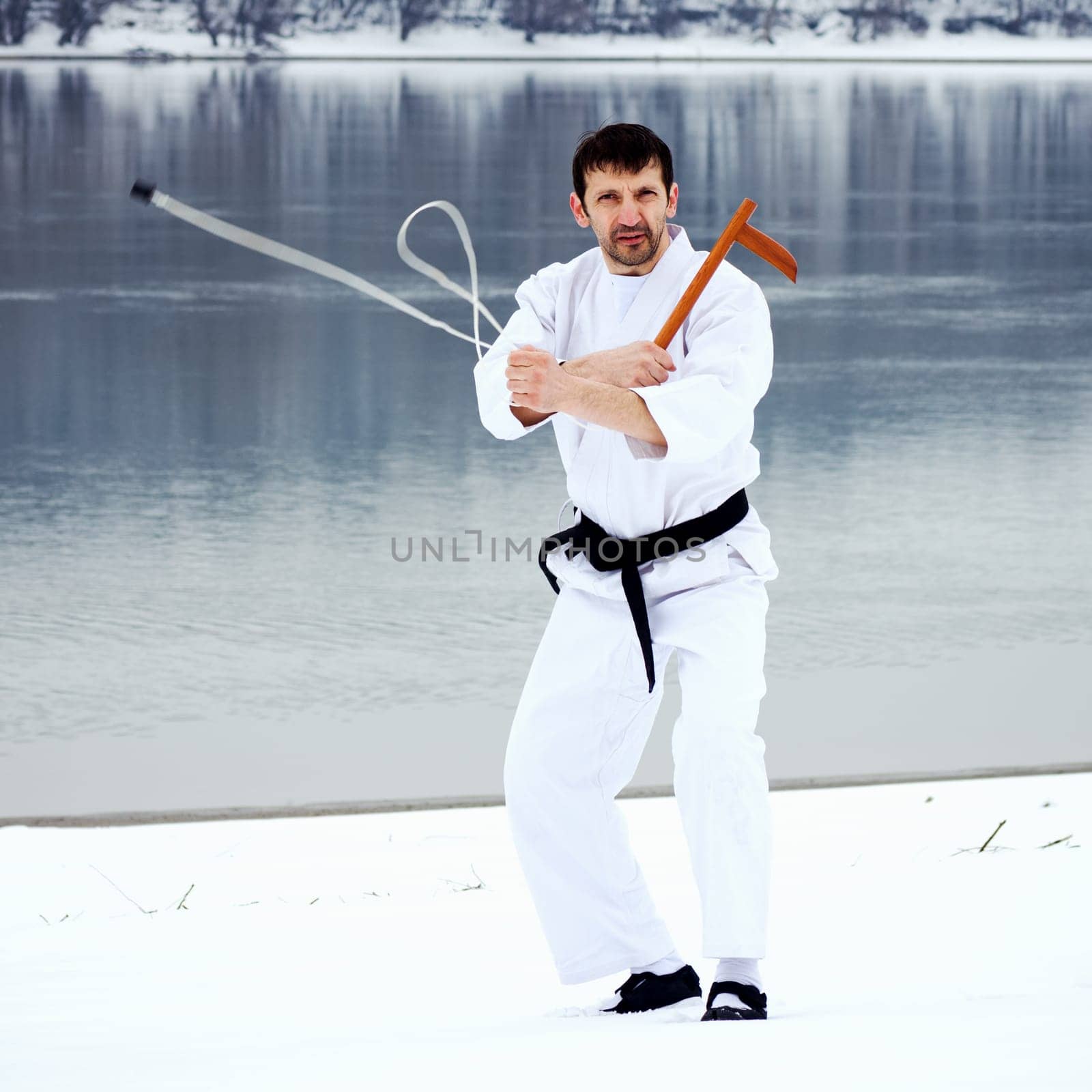 The man in white kimono and black belt is practicing using kusarigama weapon on snow at winter.