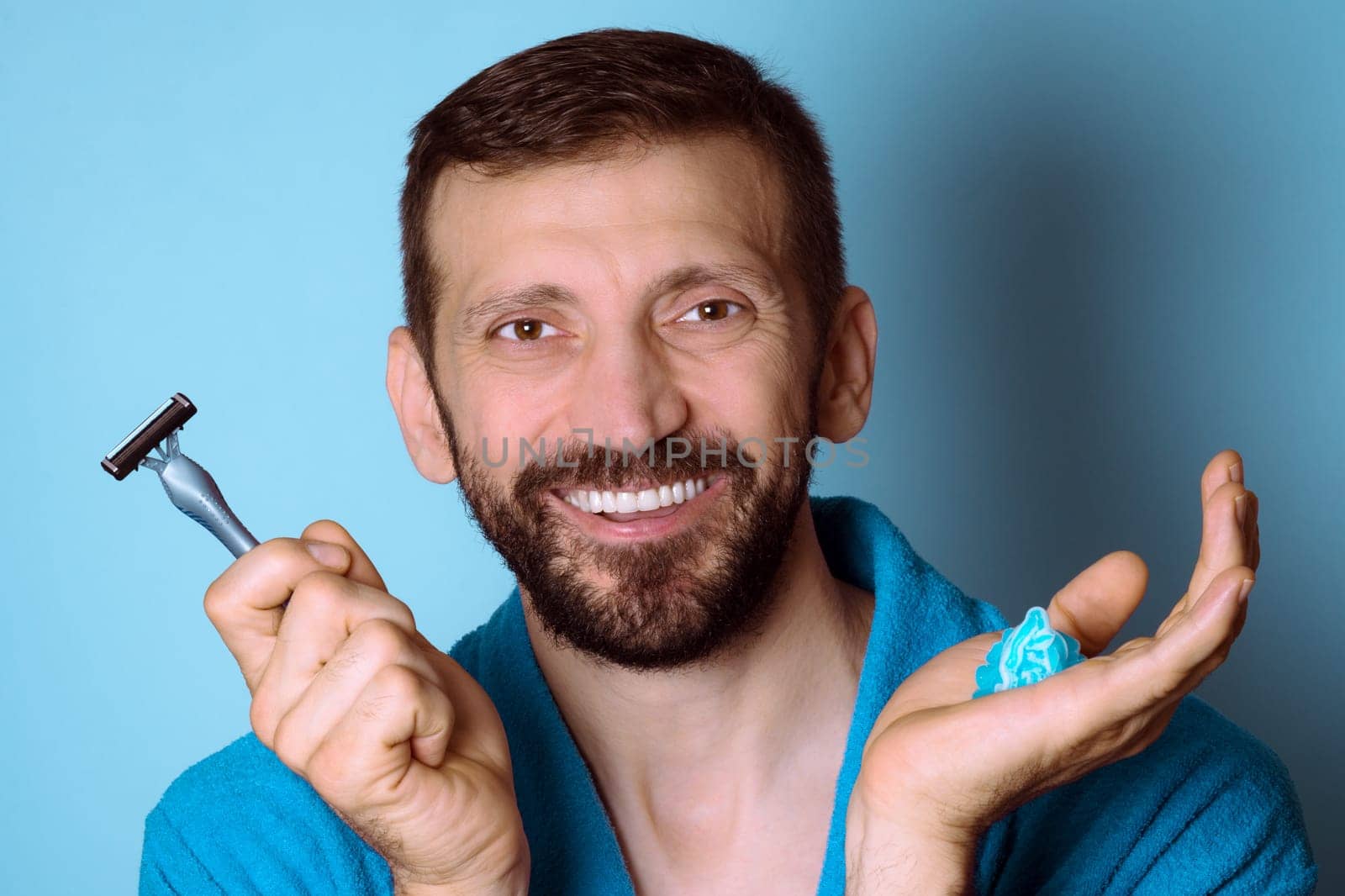The smiling bearded man is ready for shaving and holding a razor and shaving gel in hand on blue background.