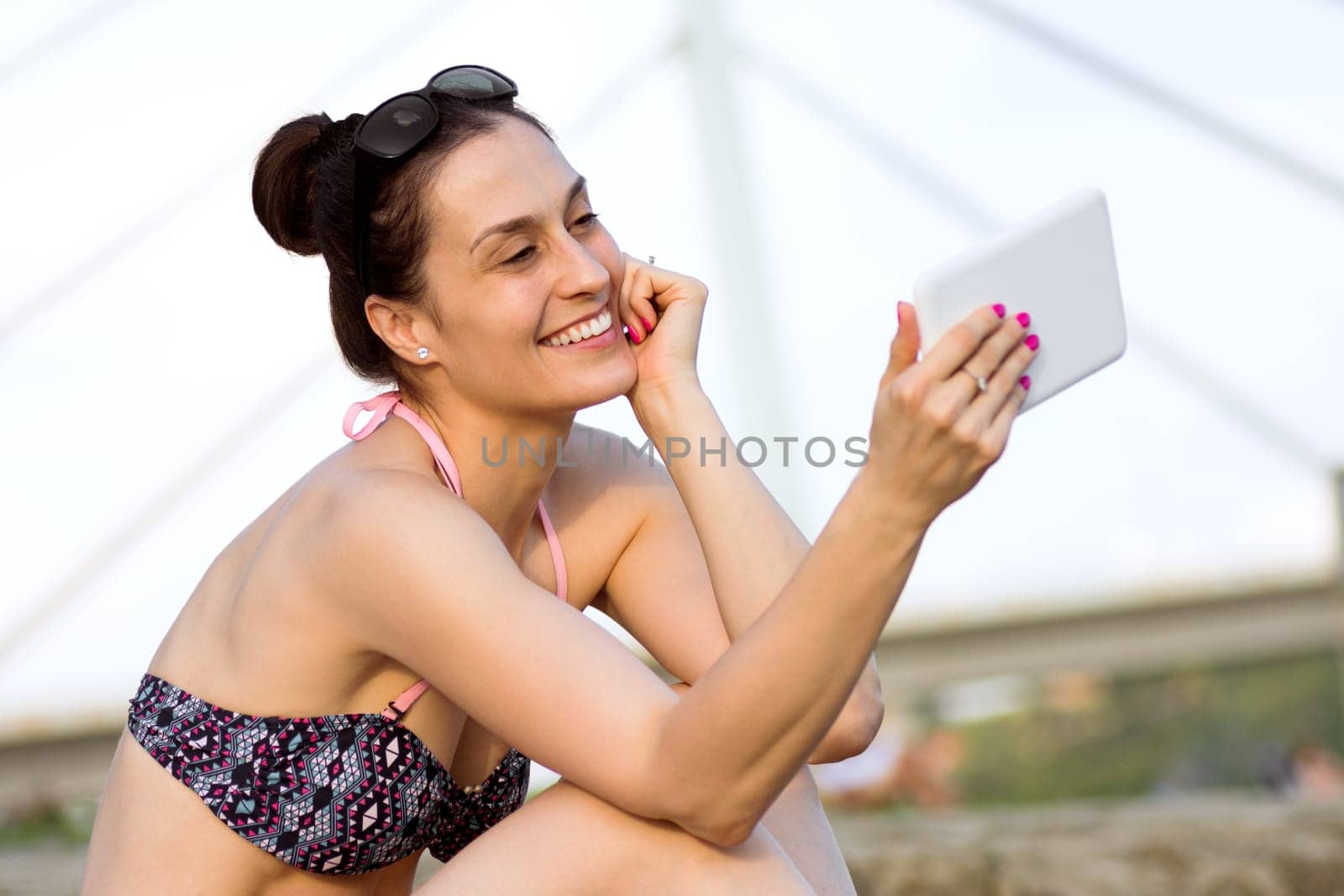 The smiling woman in bikini is using digital tablet on the beach.