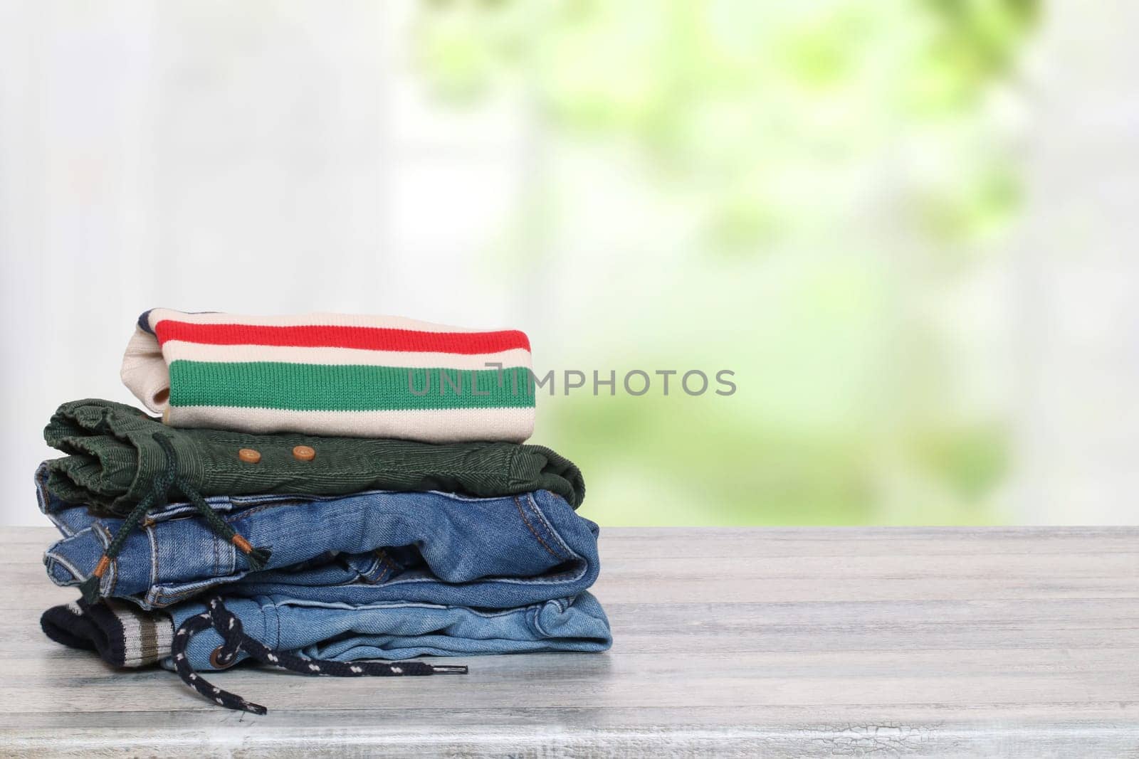 Stack of jeans. Closeup of a pile of colorful denim pants and one colorful sweater on a bright table against abstract blurred natural light green background. Space for your product display montage.