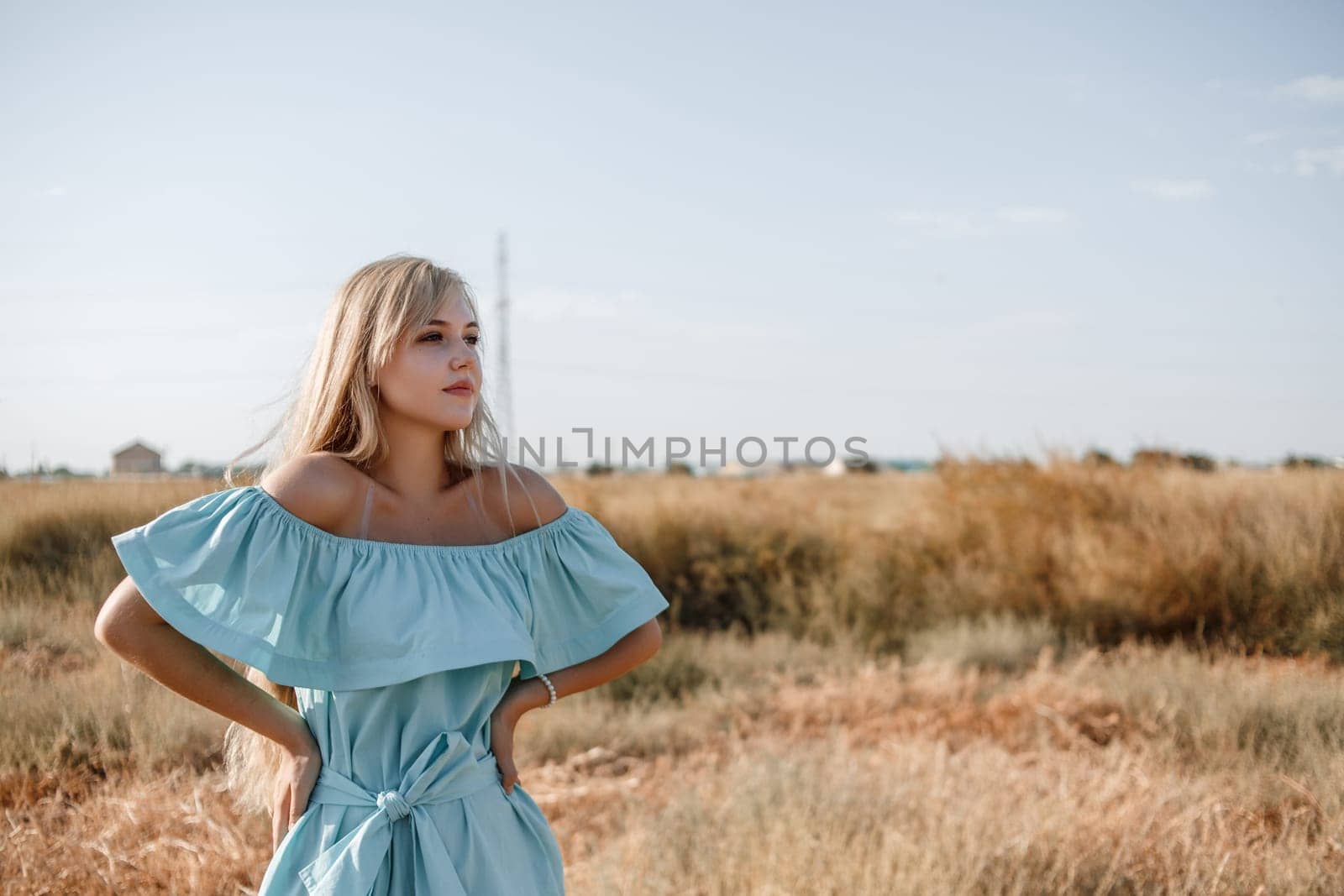 young beautiful caucasian blonde girl in light blue dress stands on the field with the sun-scorched grass