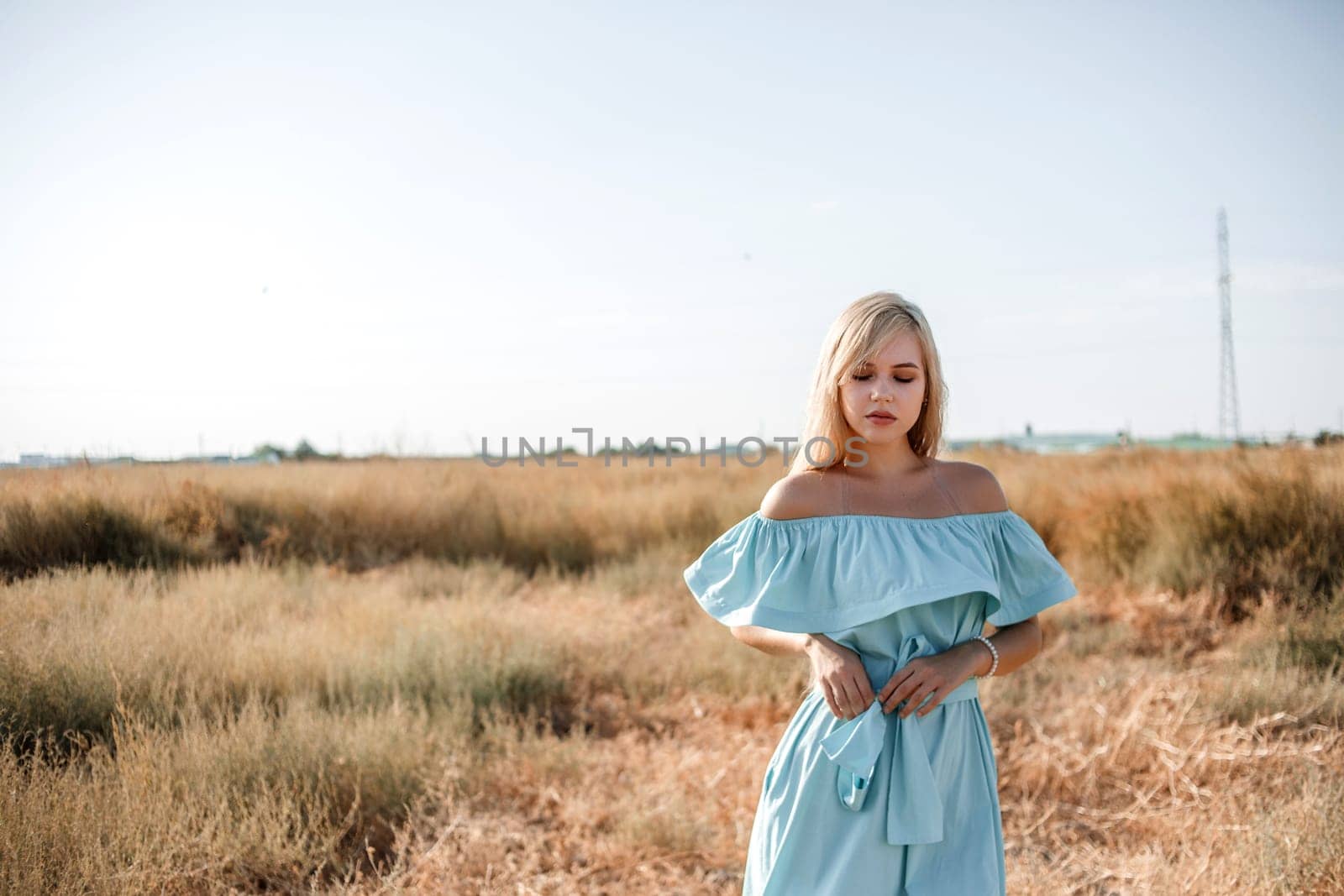 young beautiful caucasian blonde girl in light blue dress stands on the field with the sun-scorched grass
