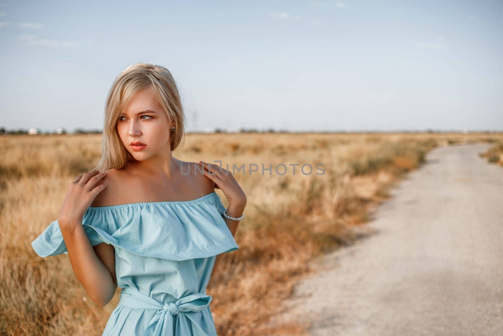 portrait of a young beautiful caucasian blonde girl in a light blue dress standing on a field with sun-dried grass next to a small country road