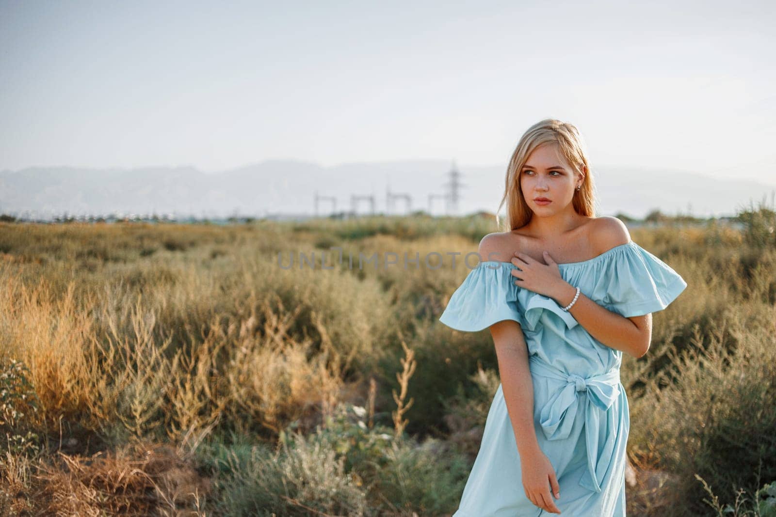 portrait of young beautiful caucasian blonde girl in light blue dress standing on the field with sun-dried grass during sunset