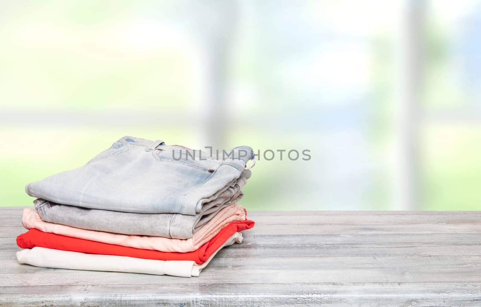 Stack of jeans. Closeup of a pile of colorful female denim pants on a bright wooden table against abstract blurred natural light pastel windows background. Space for your product display montage.