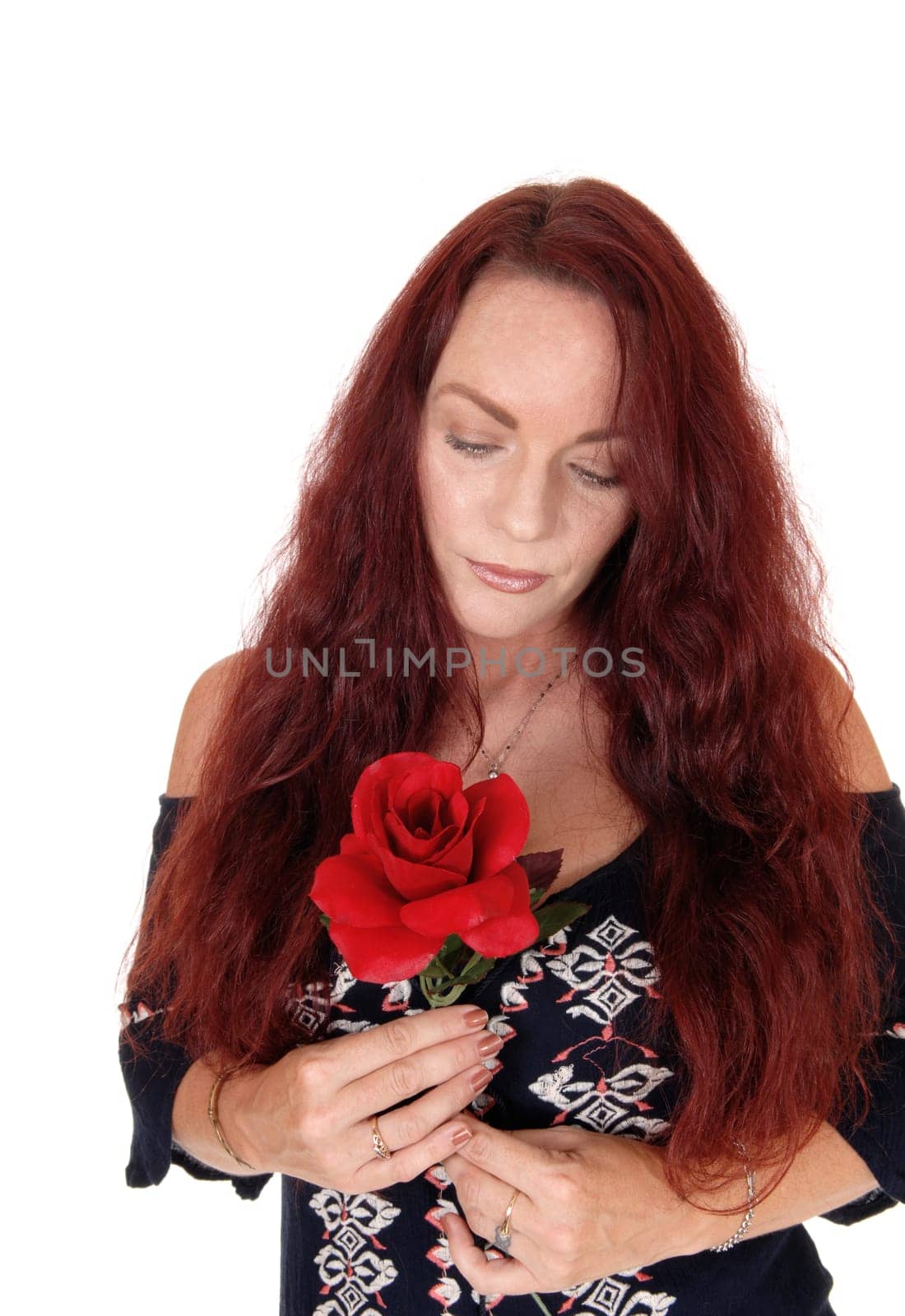 A sad looking woman with long red hair holding a red rose in her hand.looking down, standing in a black dress isolated for white background.